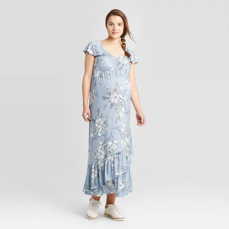 An adult wears the long light blue dress with white floral detailing, flutter sleeves and a ruffled bottom