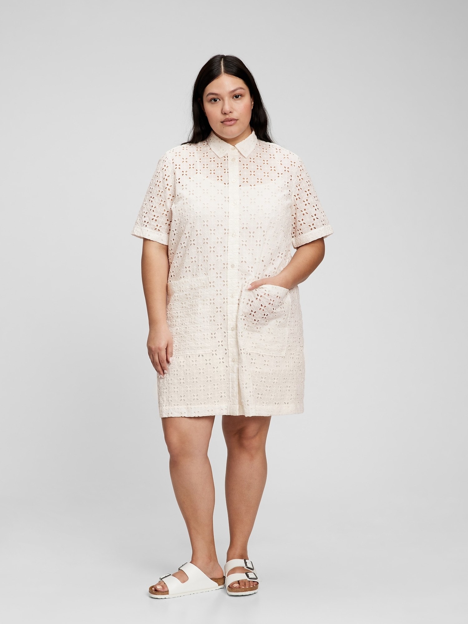 model in a white eyelet button-up mid thigh length dress with two large pockets over a white slip