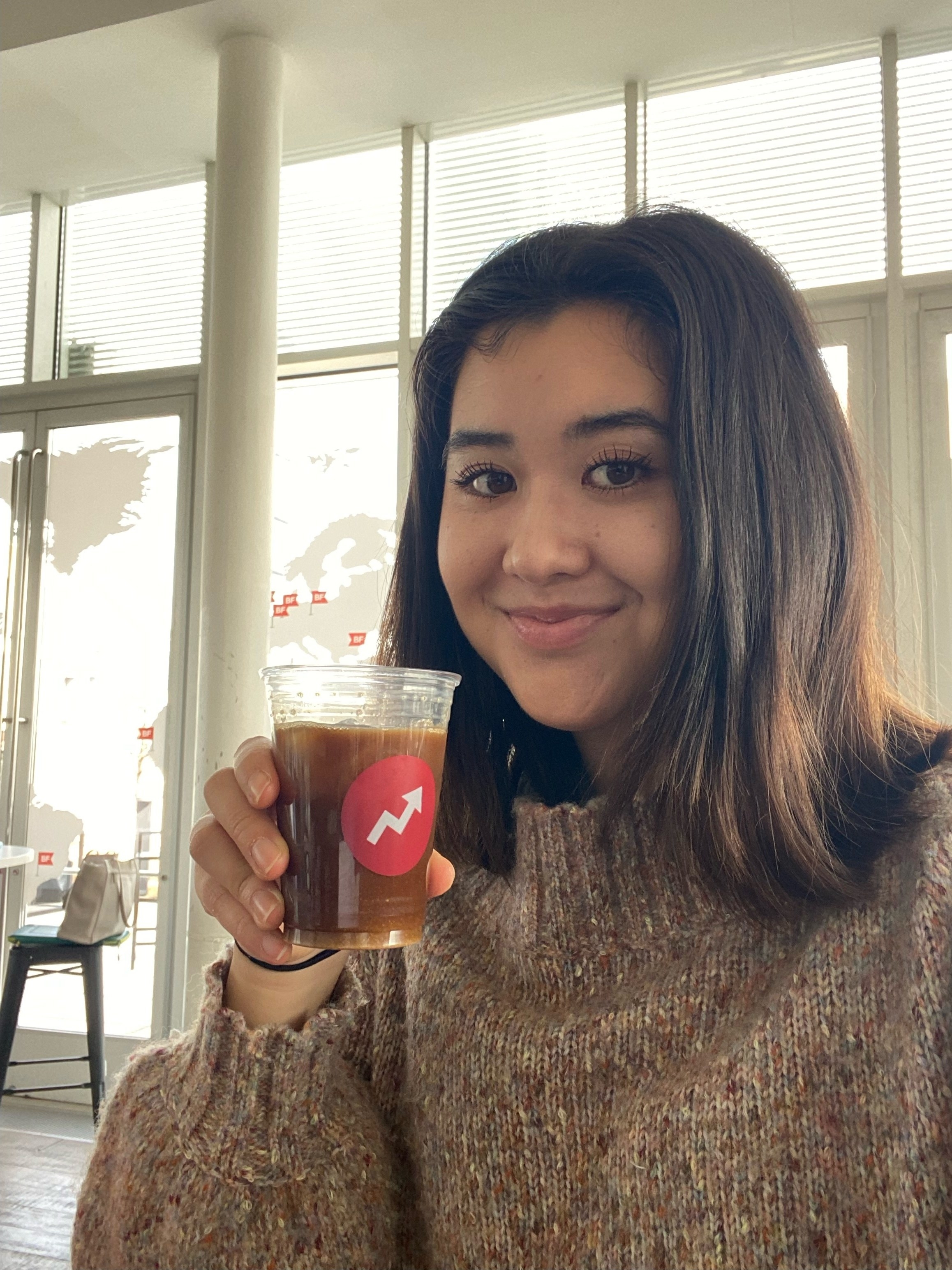 A smiling woman wearing a mock turtleneck sweater holds a plastic cup of coffee
