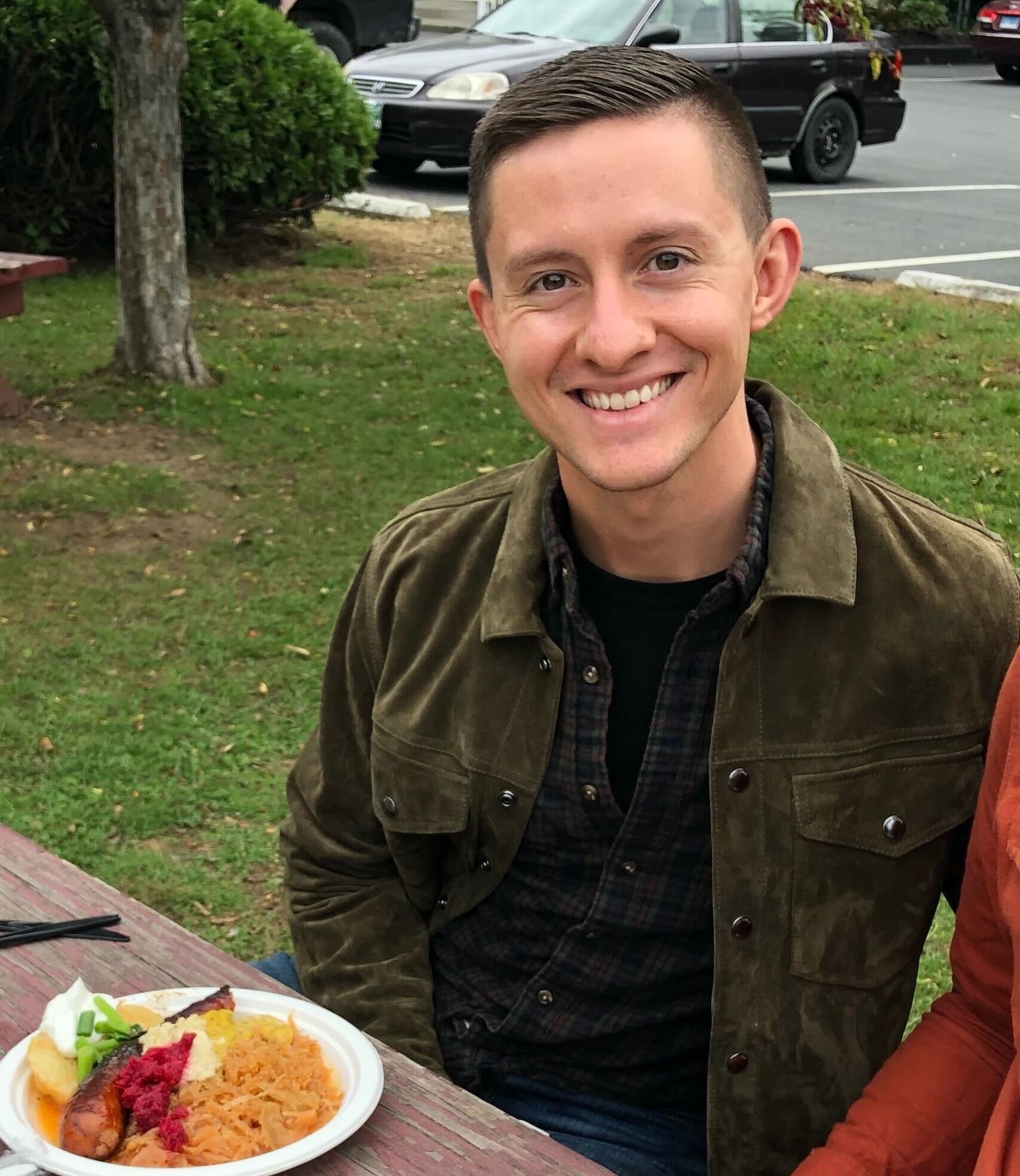 A smiling man sits outside at a table with a plate of food in front of him