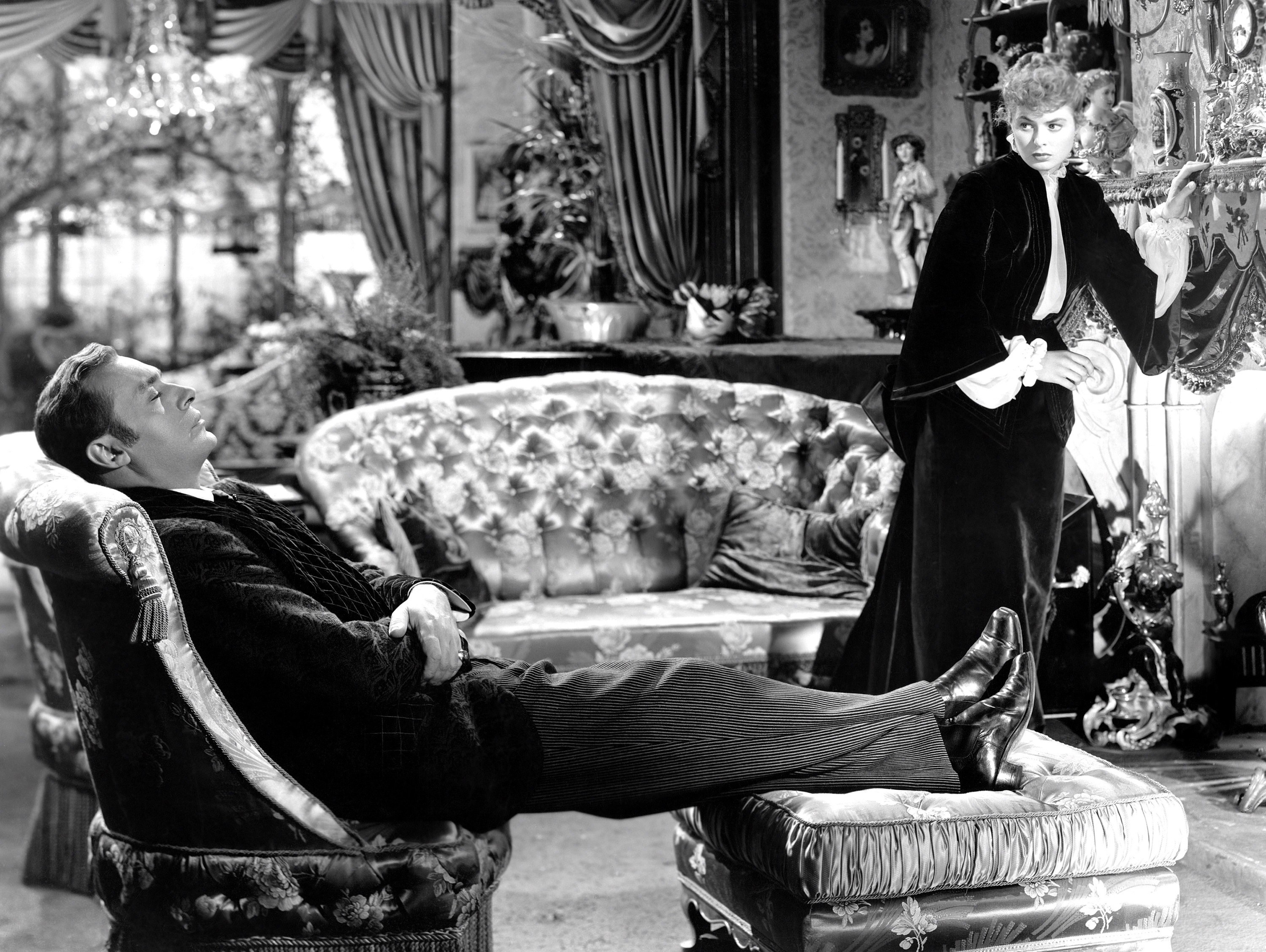 Charles Boyer sleeps in living room with his legs stretched out and resting on an Ottoman stool while Ingrid Bergman stands near the fireplace and looks at him