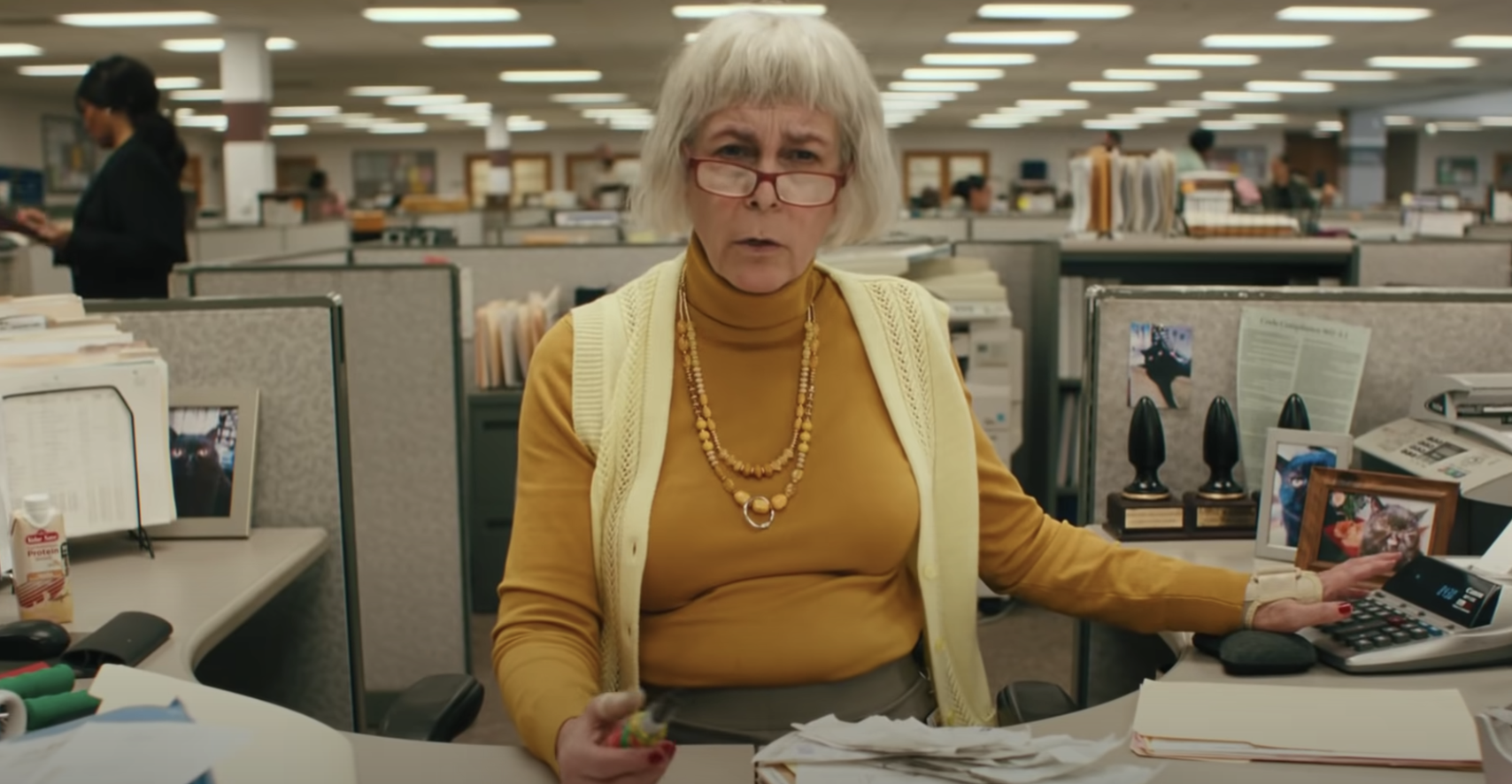 Jamie sits behind a desk in a short grey wig, opened sweater vest, not sucking in her stomach for her new role