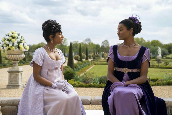 two women in regency-era attire sit on a bench with gardens behind them