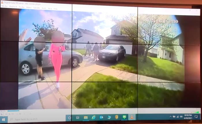 A computer screenshot of several people, one in pink, standing outside on a sidewalk with two cars nearby