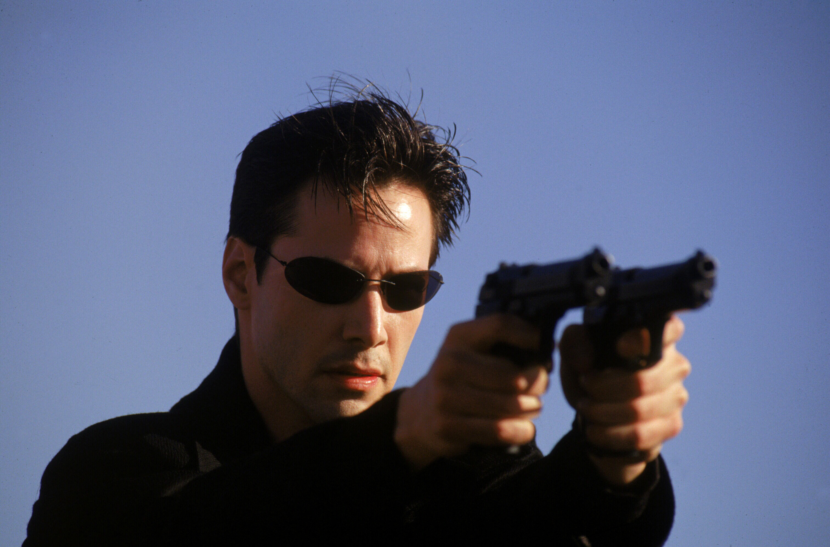 Keanu Reeves holding two guns and wearing thin sunglasses