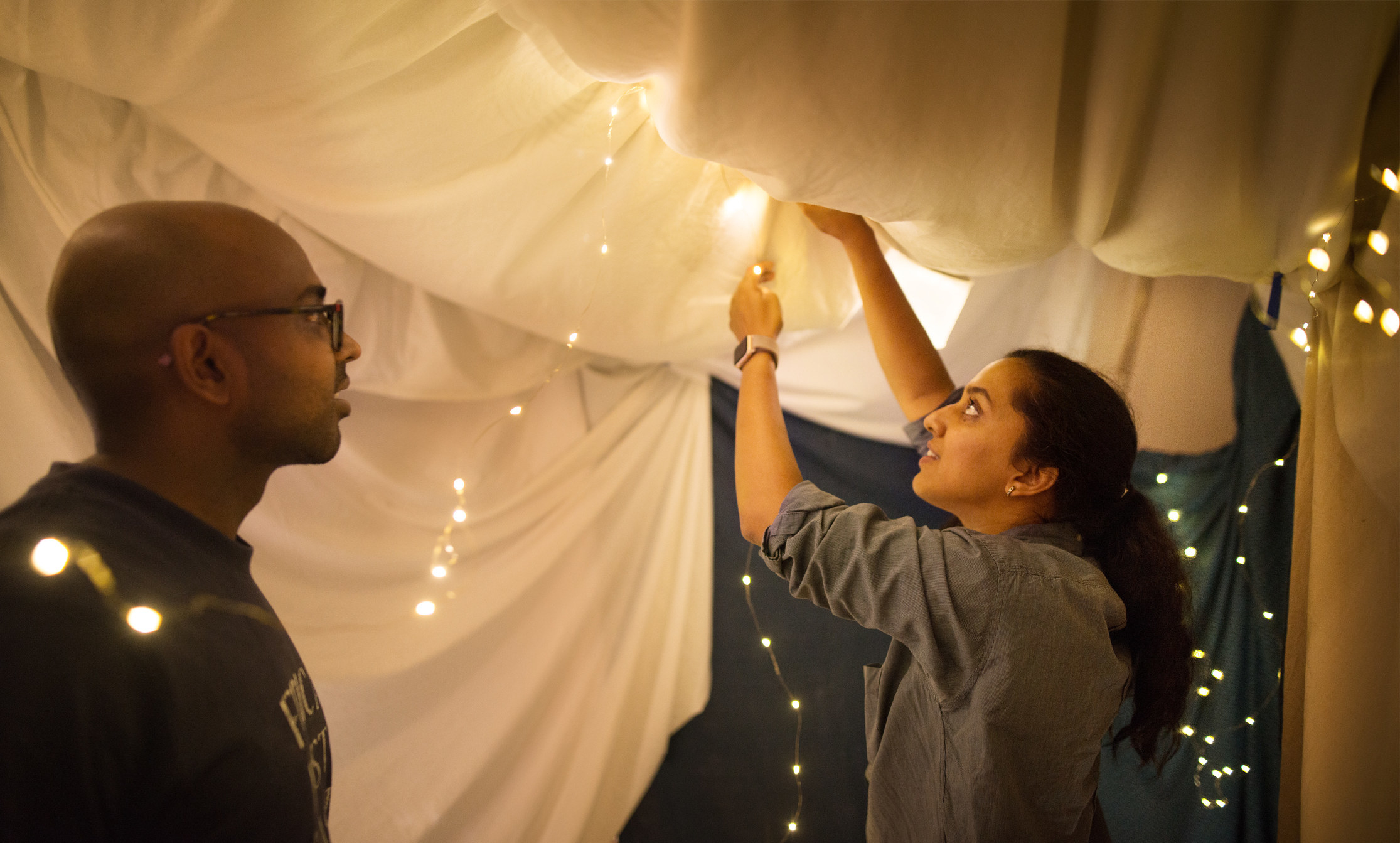 Two people drape string lights inside of a blanket fort