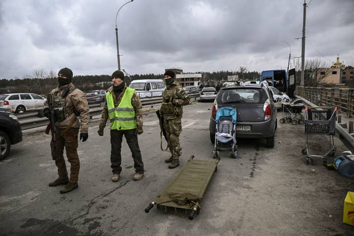Three armed men in camo stand on a bridge with cars in the background and a stretcher next to them