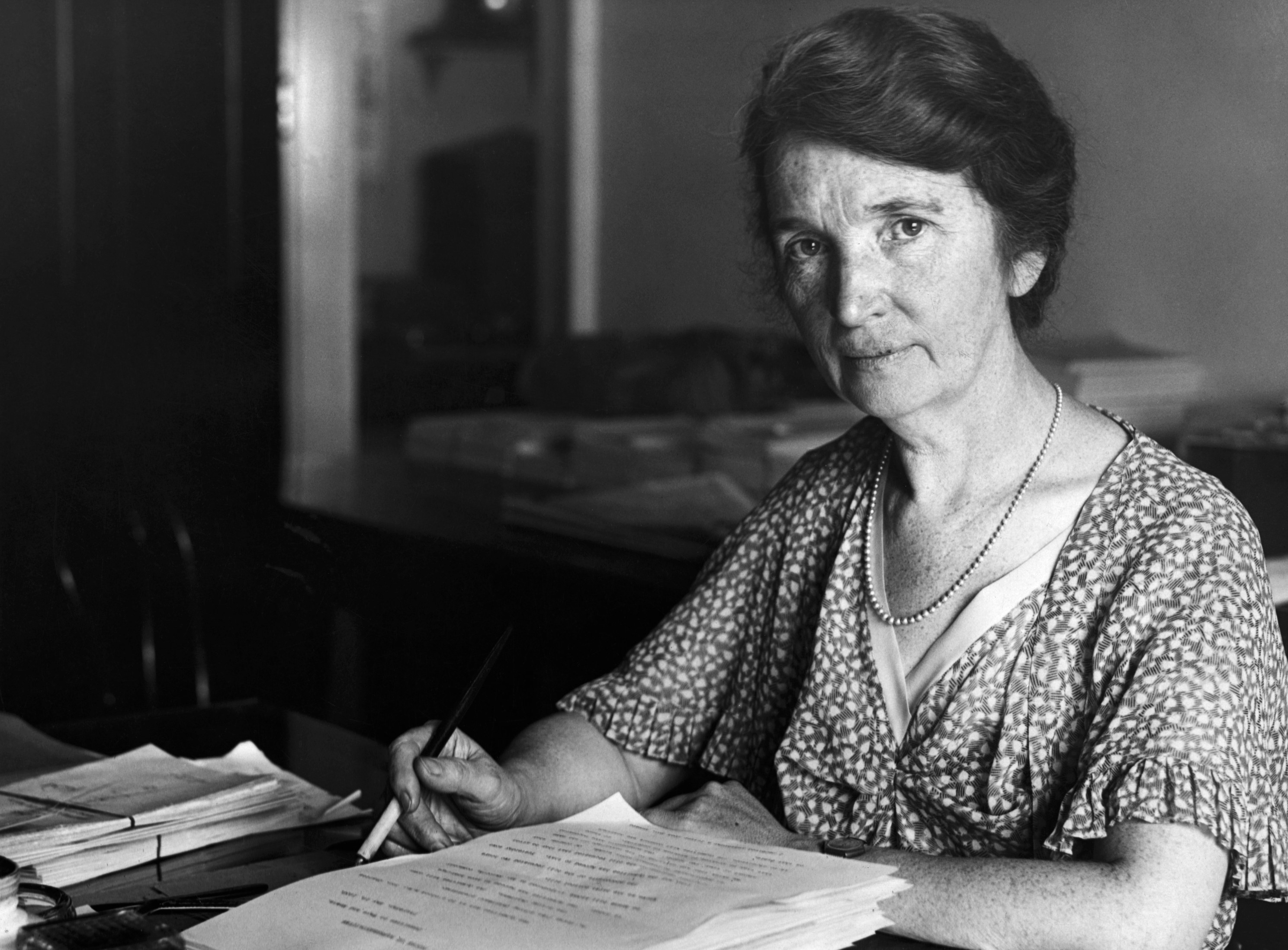 Portrait of Margaret Sanger, wearing a loose, short sleeved, patterned dress. She is looking into the camera in what looks like an office surrounding, with a stack of papers in front of her and a pen in her hand