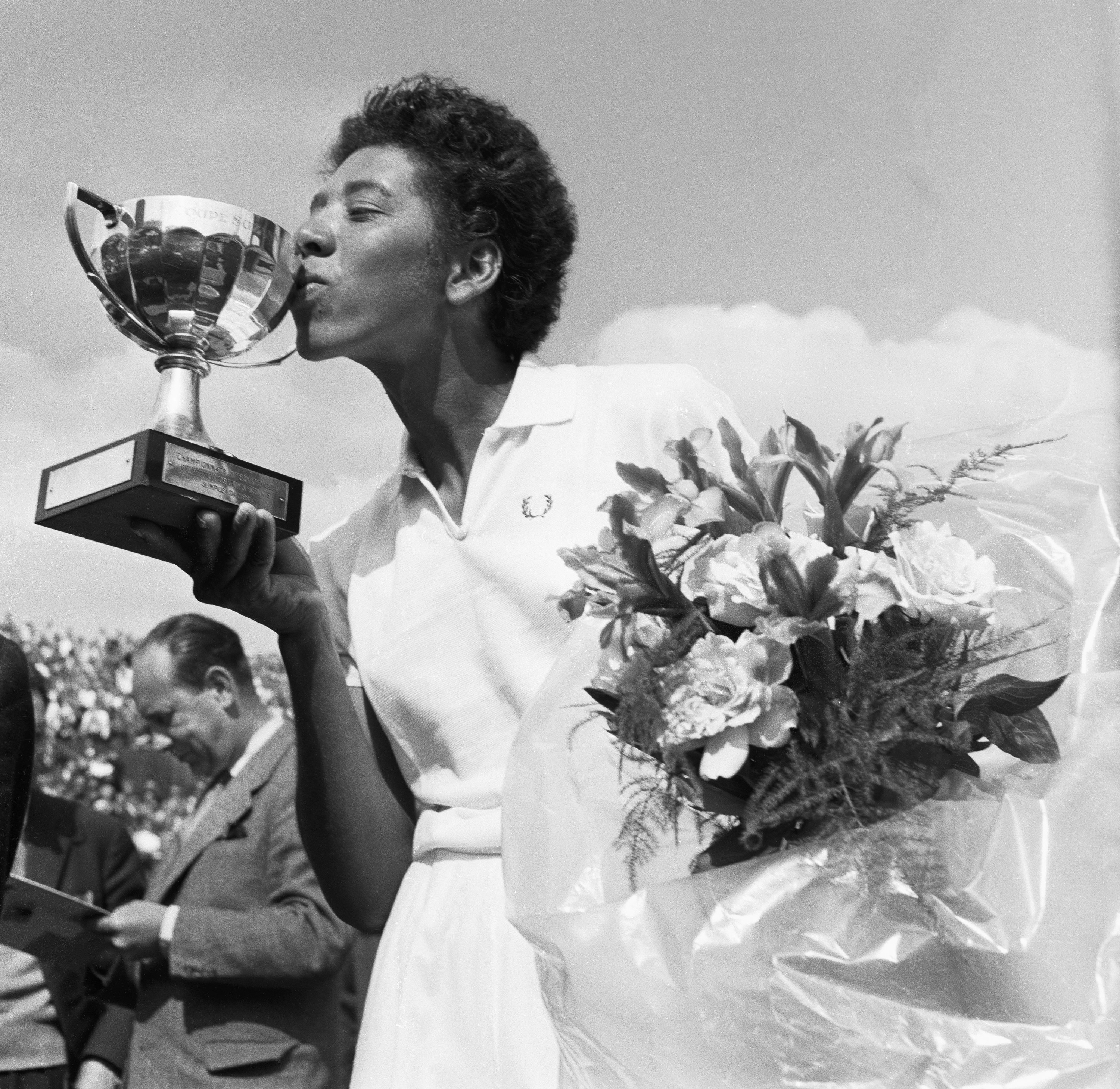Althea Gibson kissing one of her trophies and holding a bunch of flowers, dressed in her tennis whites