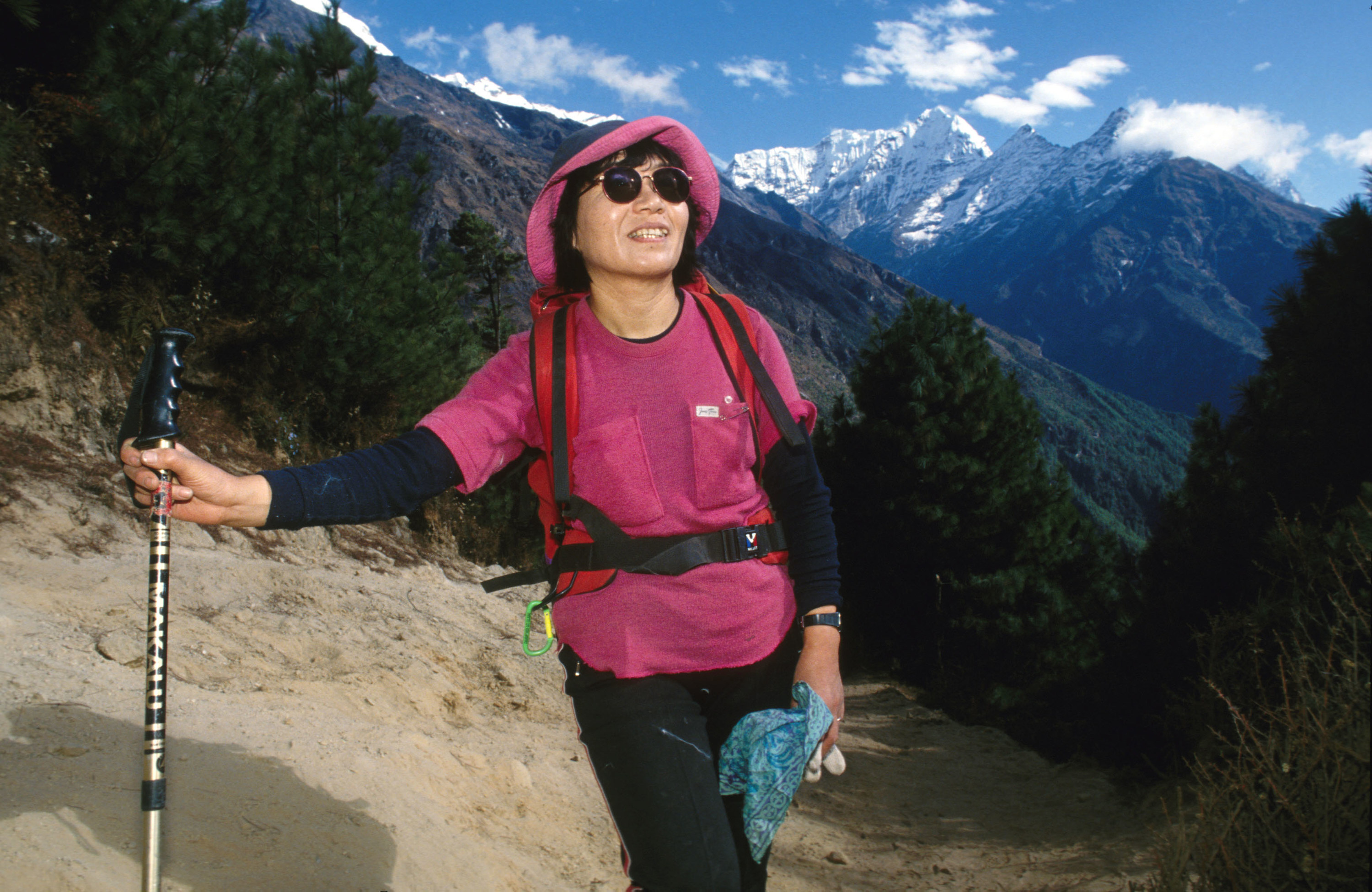 Junko Tabei wearing a backpack and holding a walking pole. She wears a hat and sunglasses and smiles as she looks ahead, a mountain scape behind her