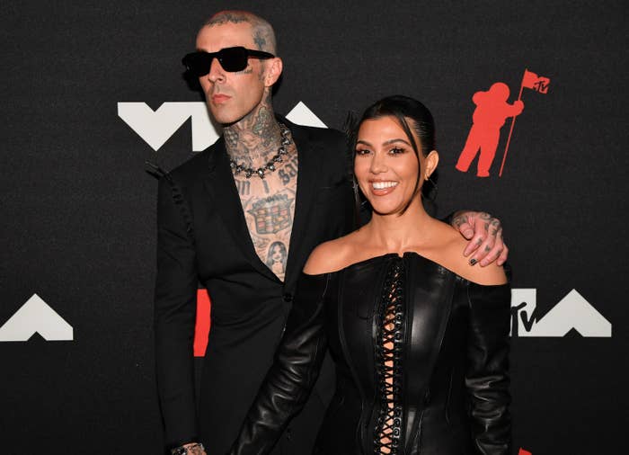 Travis Barker in a suit without a shirt and Kourtney Kardashian in a leather lace-up dress at an event