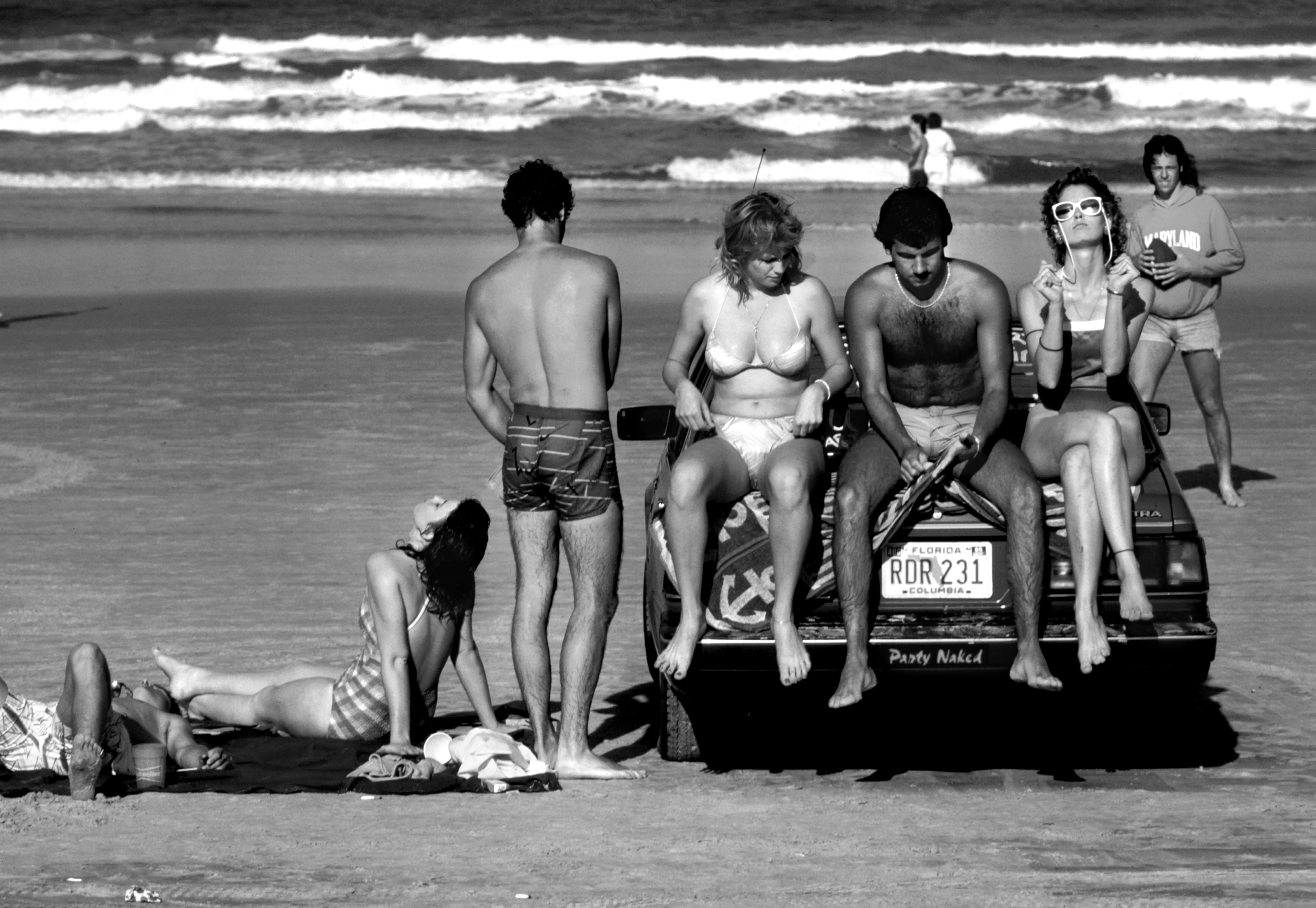 three people sitting on the back of a car with other beachgoers around them and the ocean in the background 