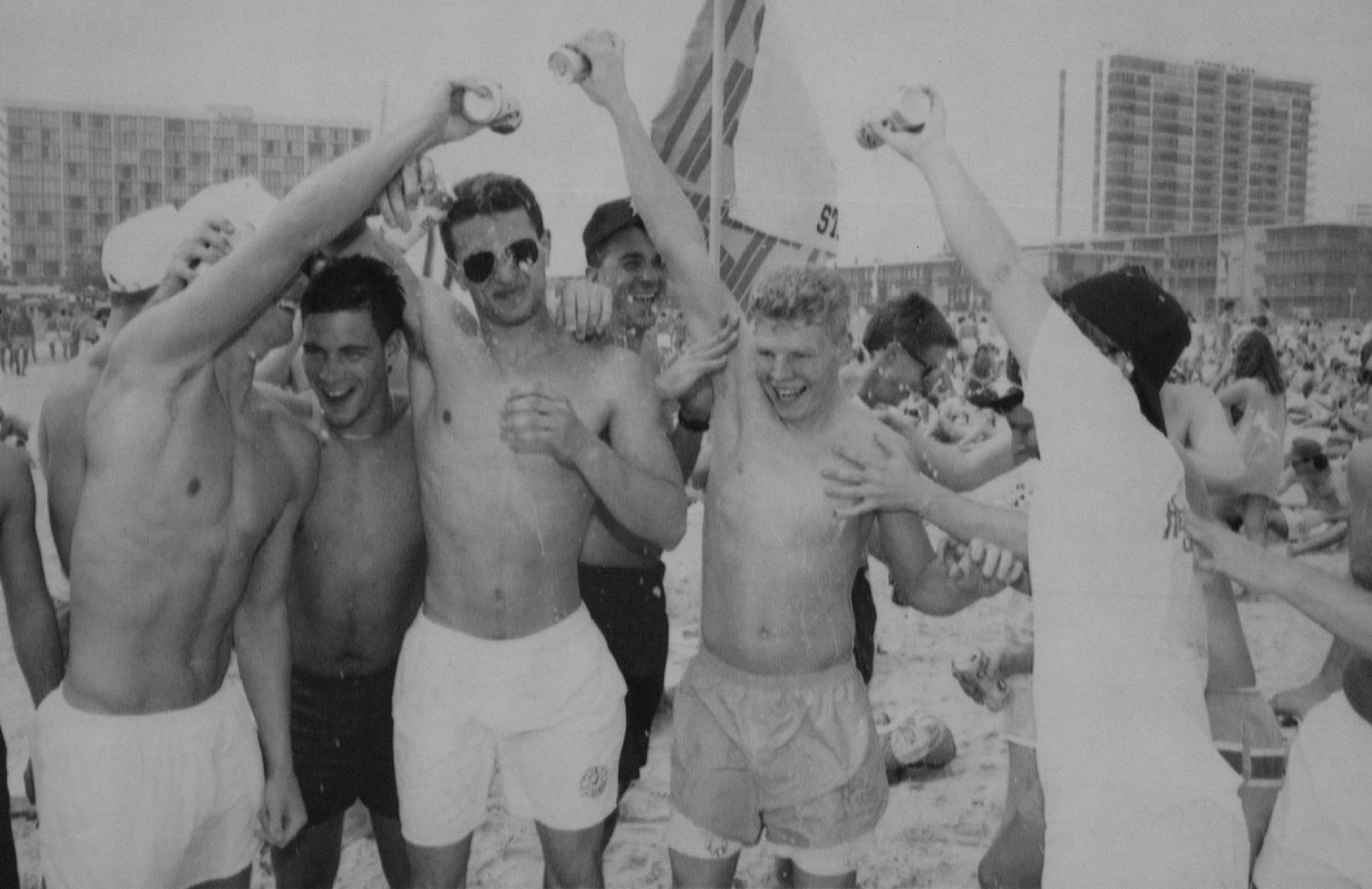 a group of shirtless young men pour beer all over each other on a beach with buildings behind them 