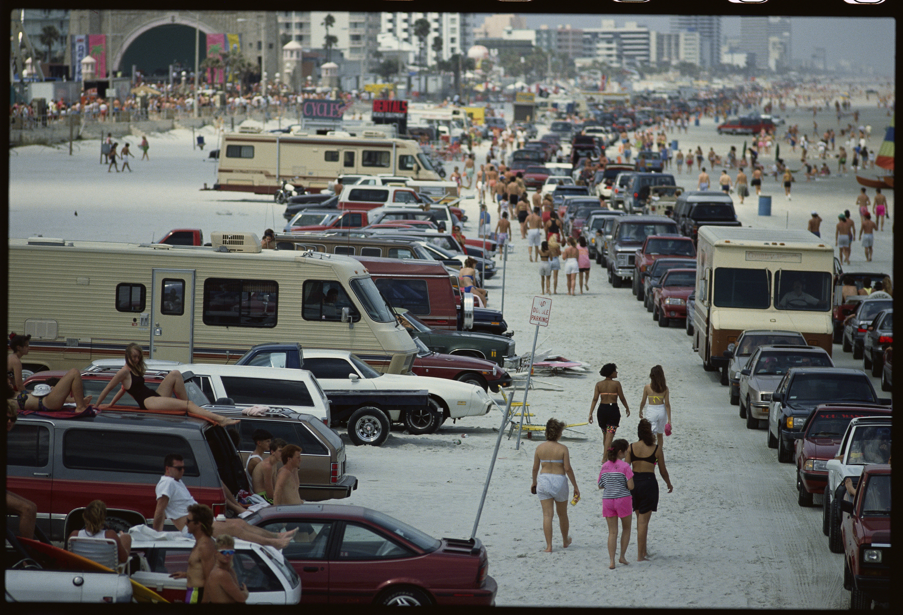 a group of people walk in between rows of cars with people sitting on car roofs on a beach 