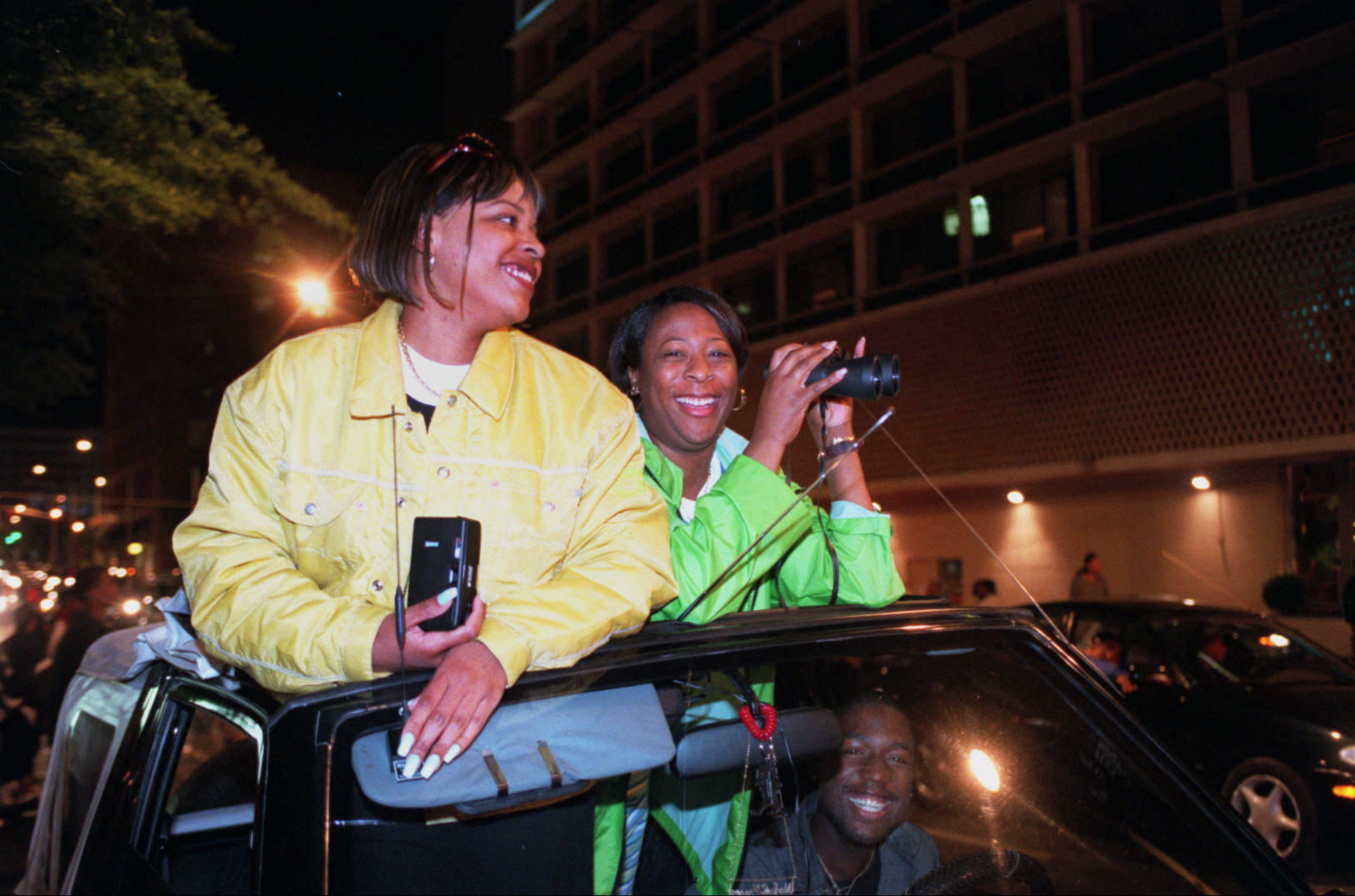 Two people with cameras poke their heads out of the sunroof of a car at night, smiling for the camera