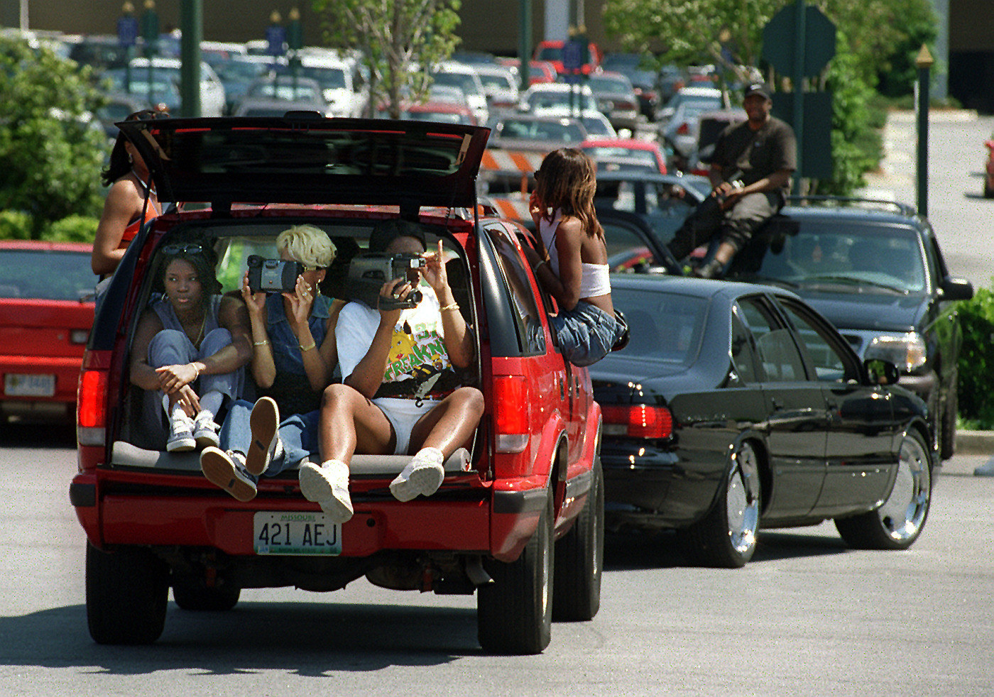three women sit in the back of a car with the trunk open and videotape the scene, two other women are hanging out the windows