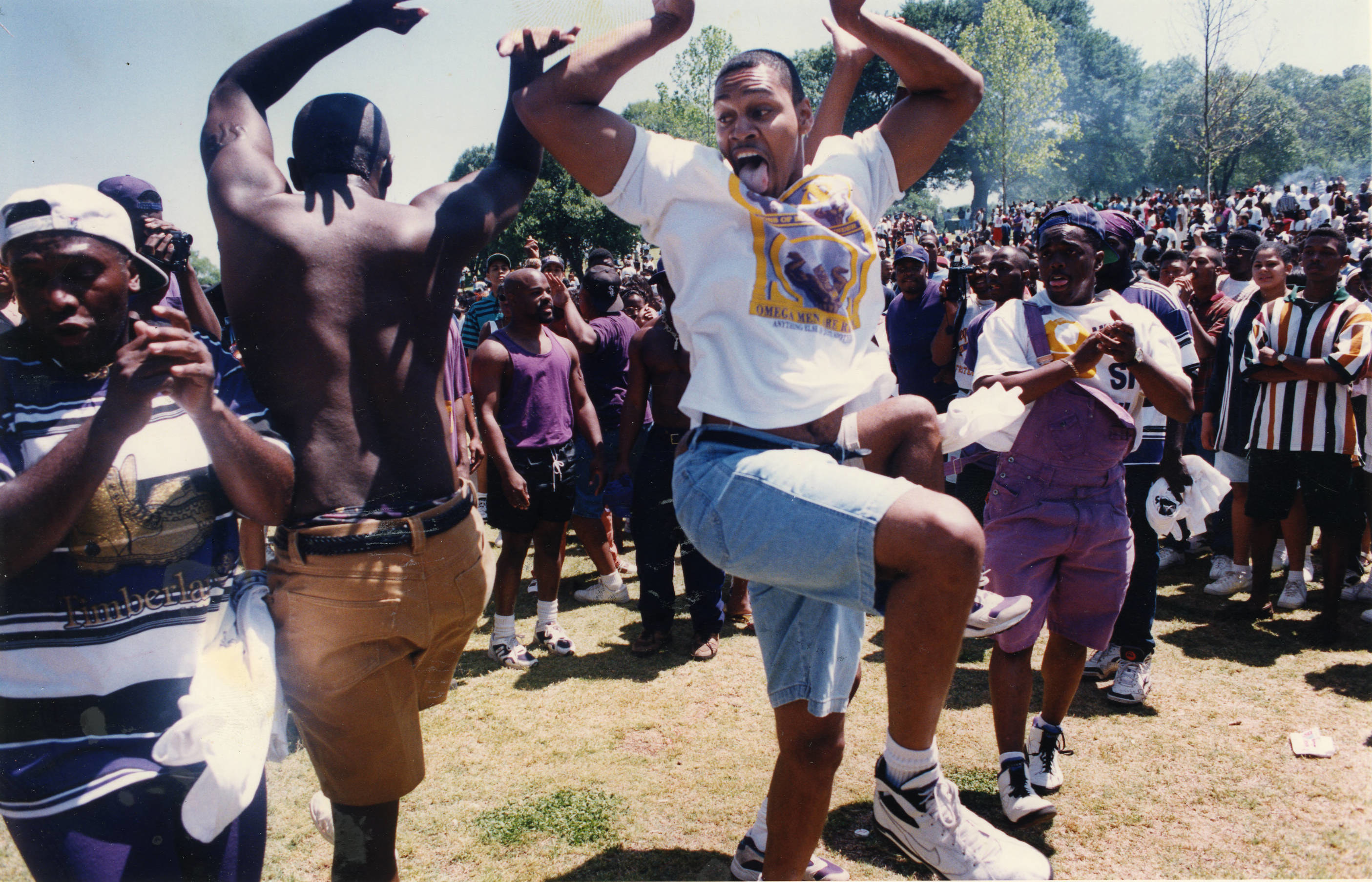 people dance in a group on grass at a park with a large crowd watching 