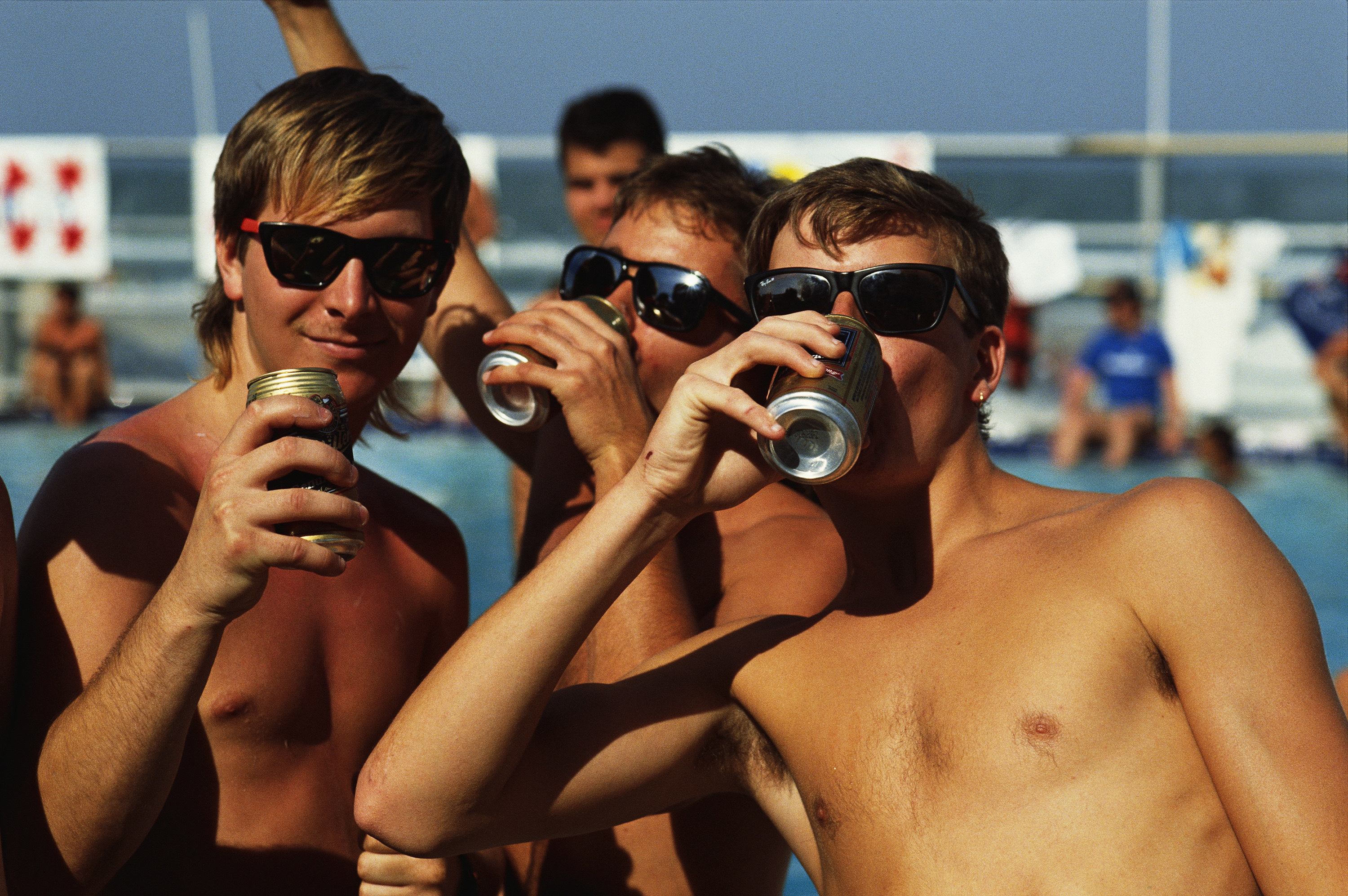 three young men in sunglasses and with no shirts drink beer in front of a pool