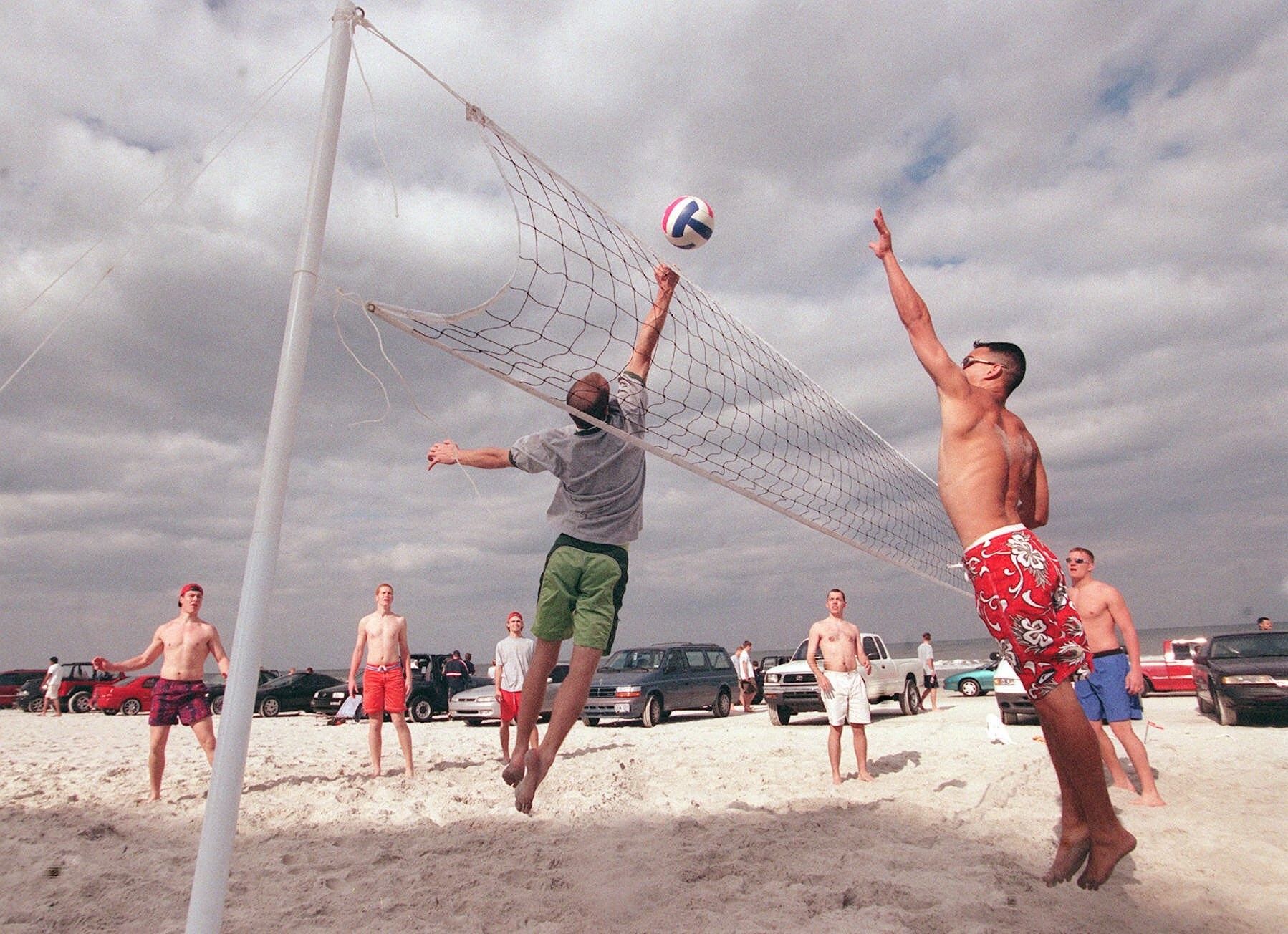 young men play volleyball on the beach with a row of cars behind them 