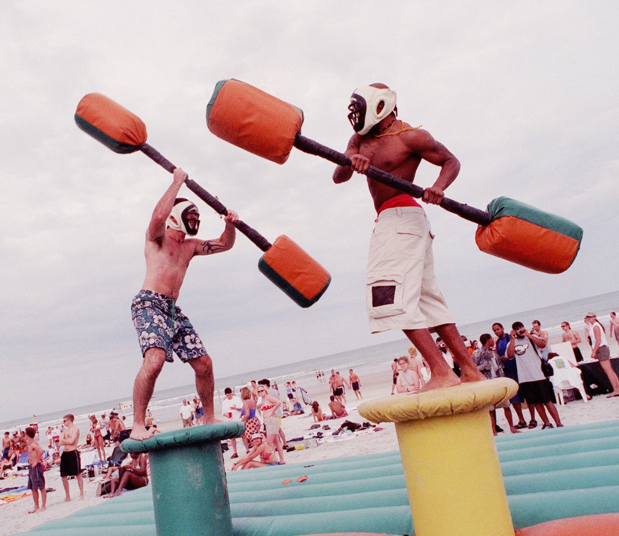 two men on podiums try to knock each other off with giant inflatable q tips 