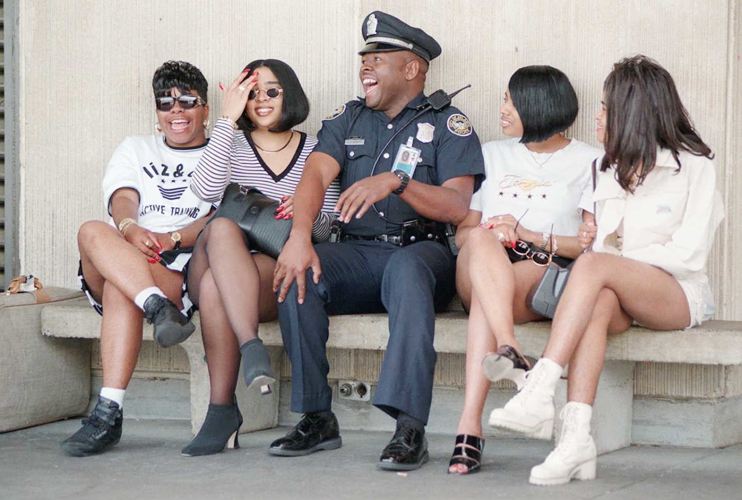 four women in shorts sit on a bench and laugh with a uniformed policeman in the middle 