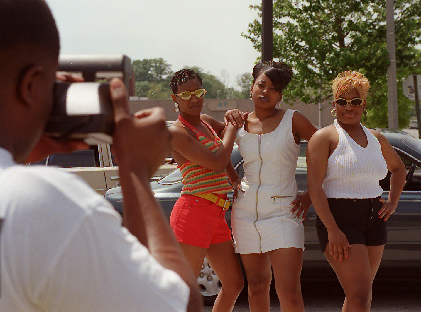 three women pose in front of a car and a man takes a photo in the foreground 