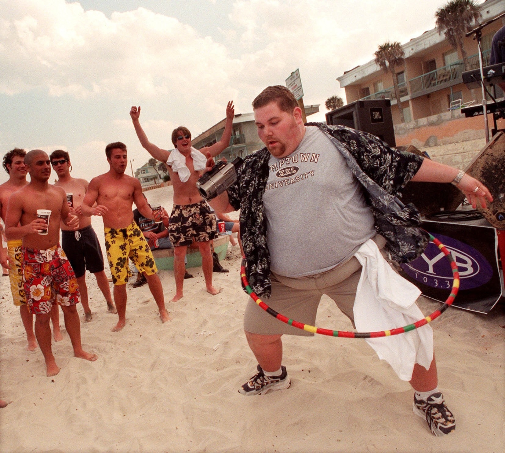 A man hula hoops in the foreground while a group of shirtless young men in board shorts cheer him on with buildings in the background