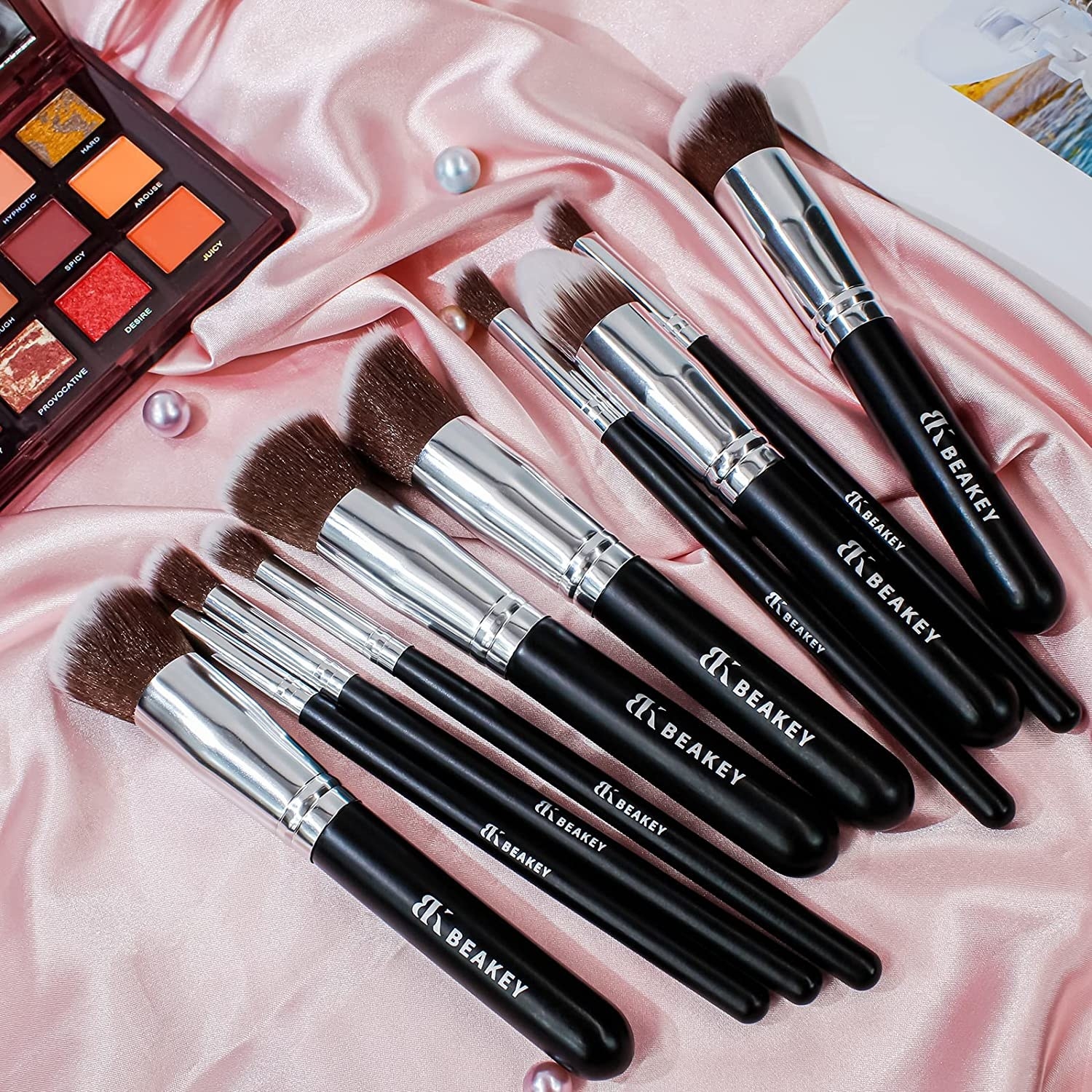 The brushes on a satiny surface next to an eyeshadow palette and some loose pearls