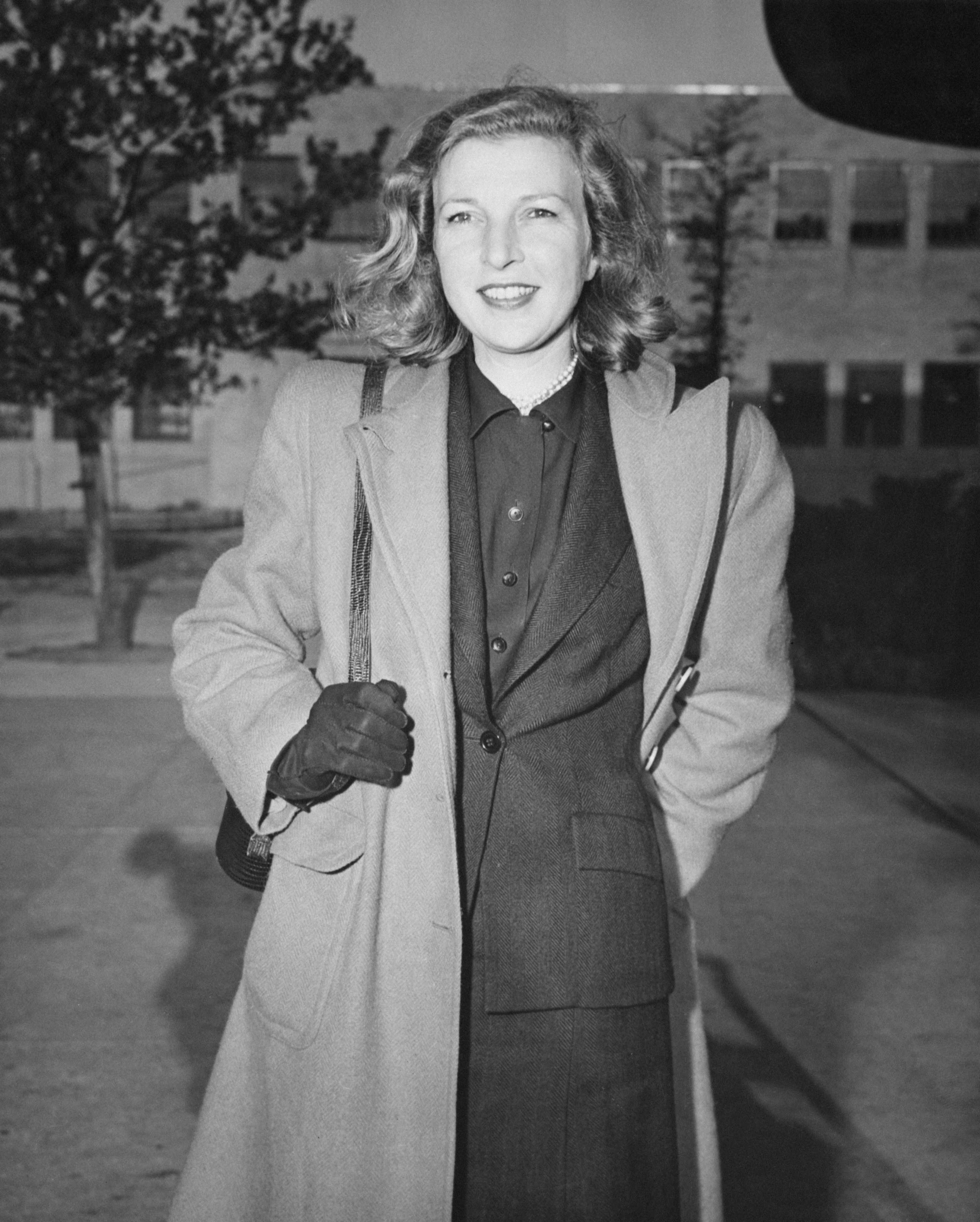Martha Gellhorn wearing a three piece suit with a trenchcoat over it, holding her bag strap in one gloved hand. She has short wavy hair and is smiling