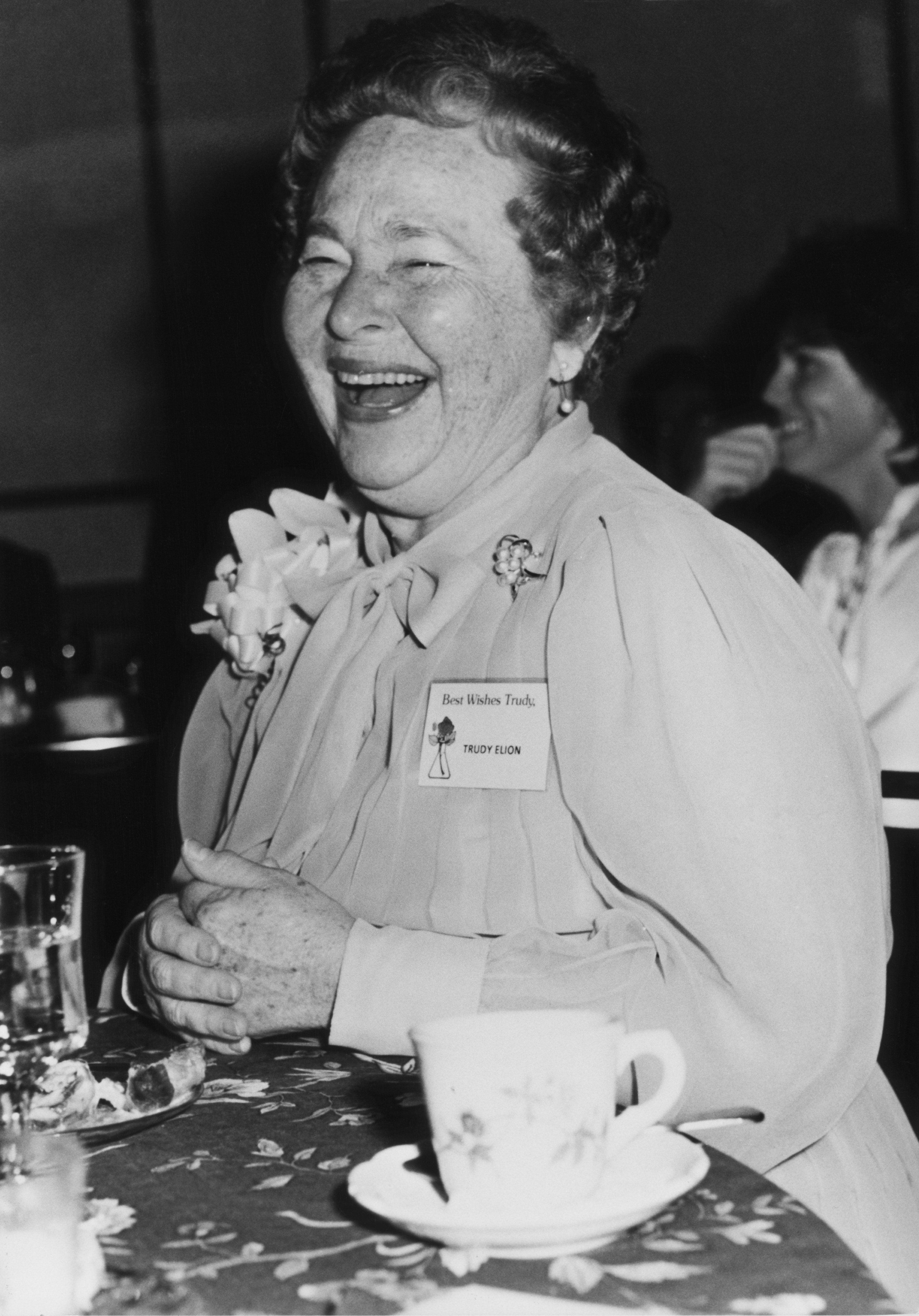Gertrude Elion laughing, sitting at a table with a floral tablecloth and a teacup and saucer in front of her