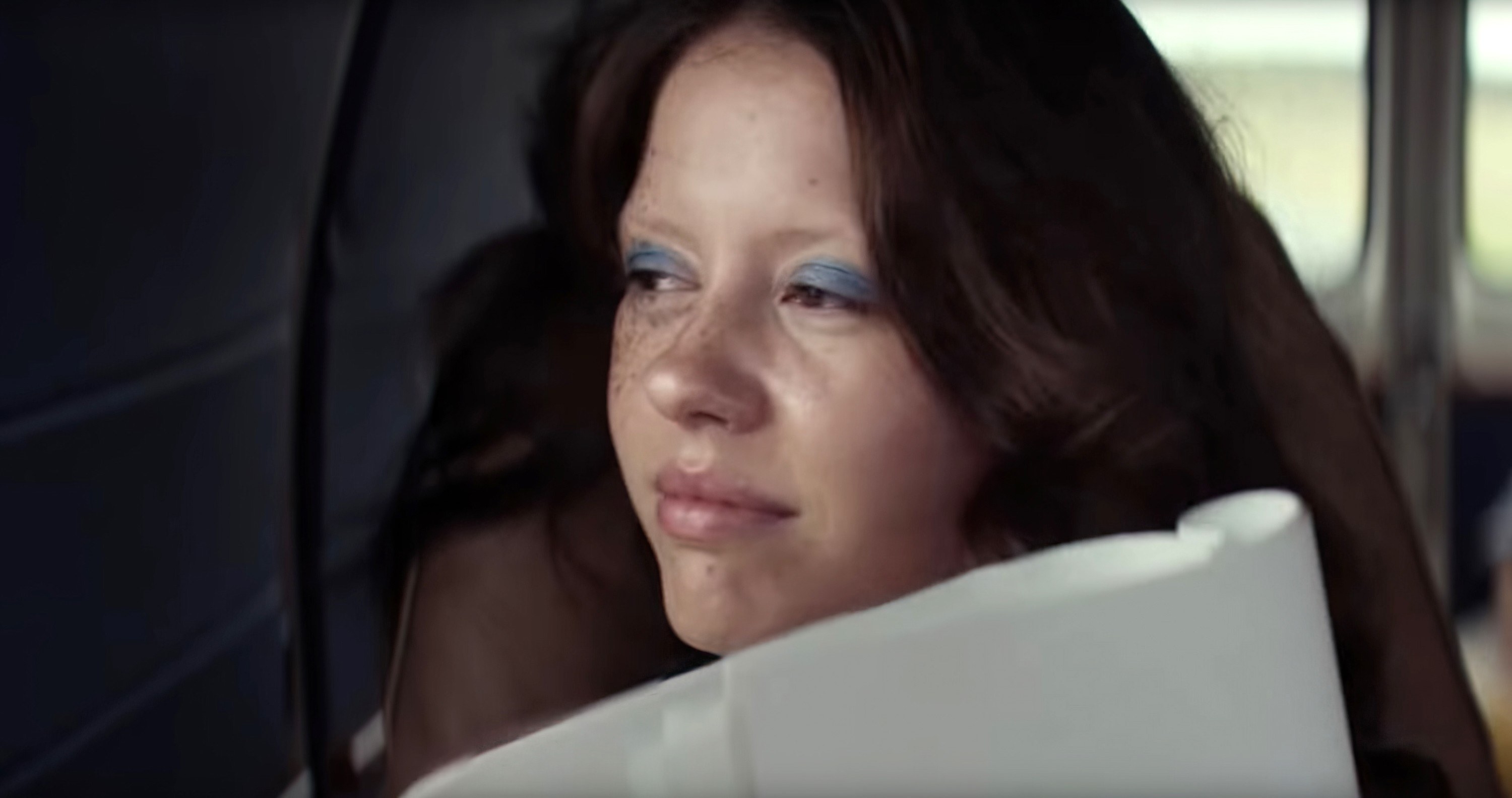 Mia Goth in her blue eyeshadow, reading a script, looking out the window as she rides in the passenger seat of a van