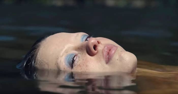 Mia Goth as Maxine, wearing bright blue eye shadow, swimming in a lake, head above water