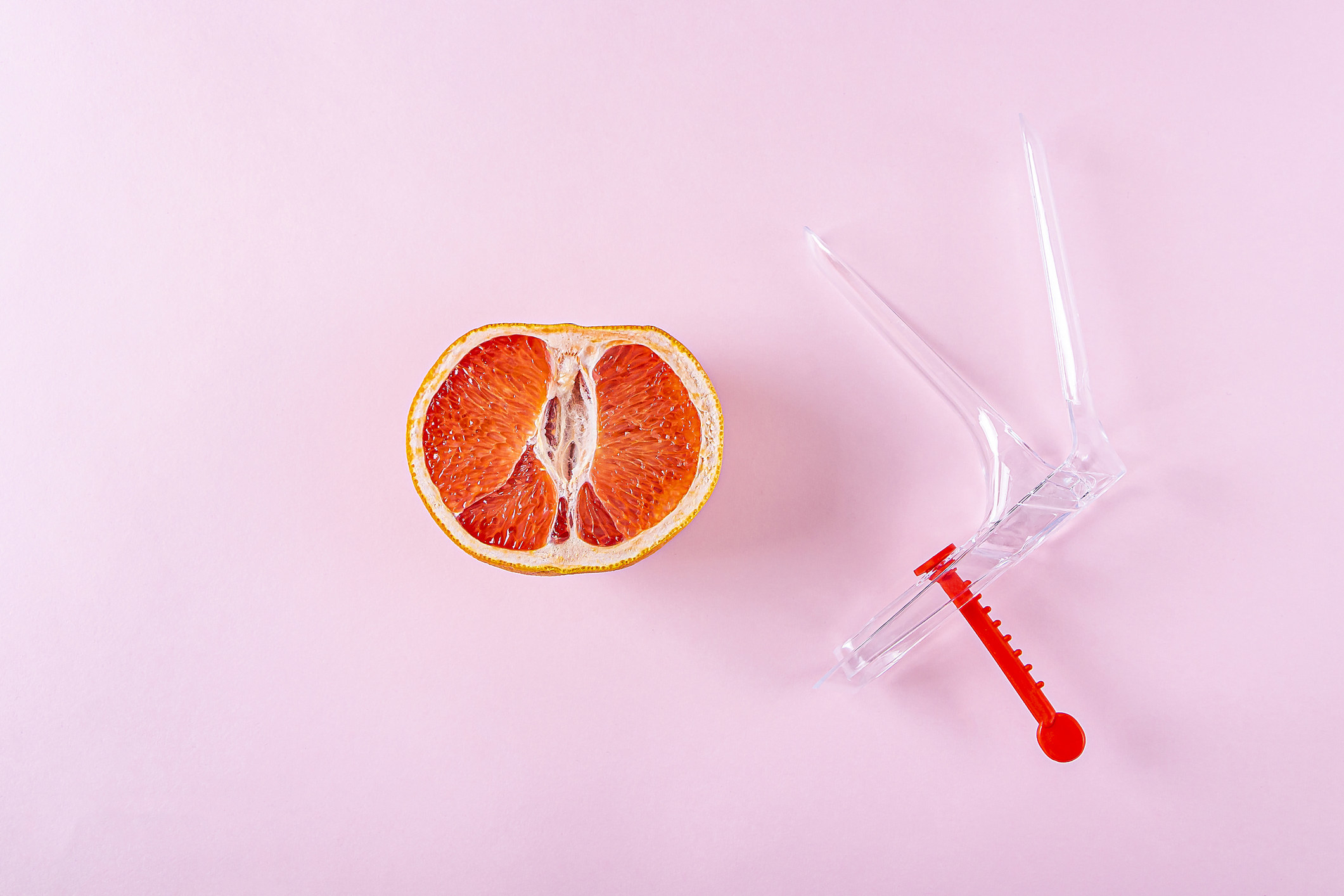 A piece of fruit depicting a vagina on a pink background color next to a gynecological speculum