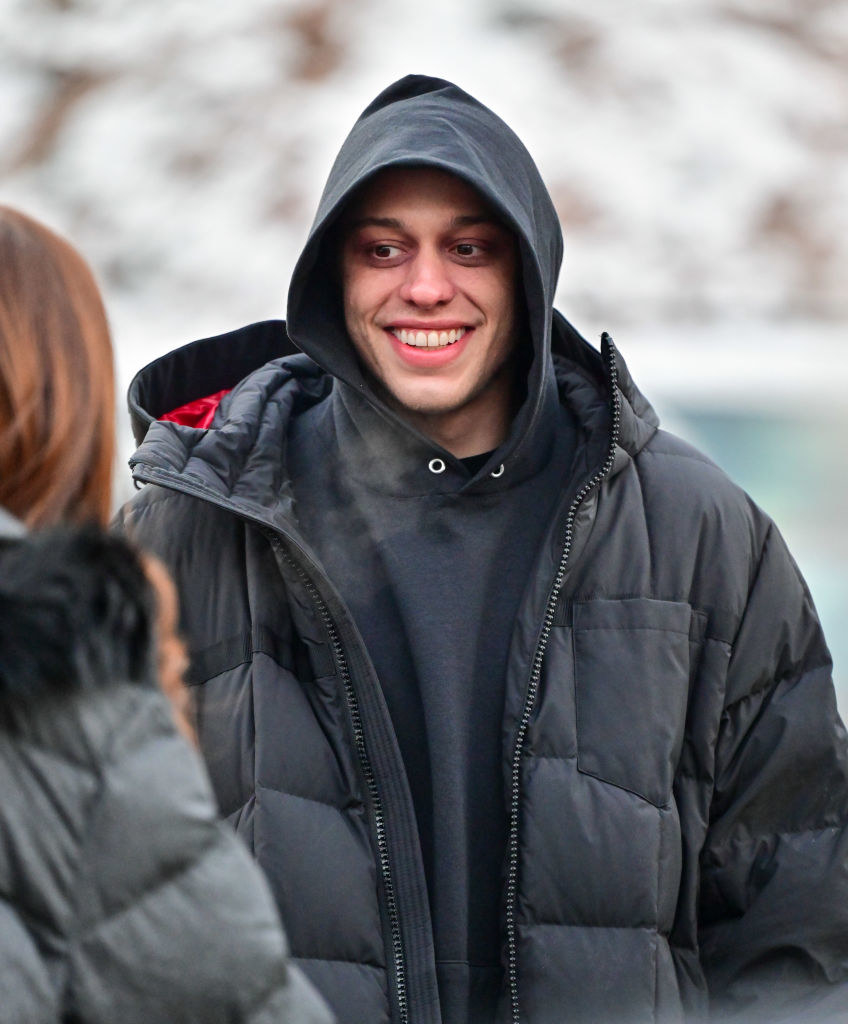 Pete Davidson, wearing a puffy coat and a hoodie, smiles