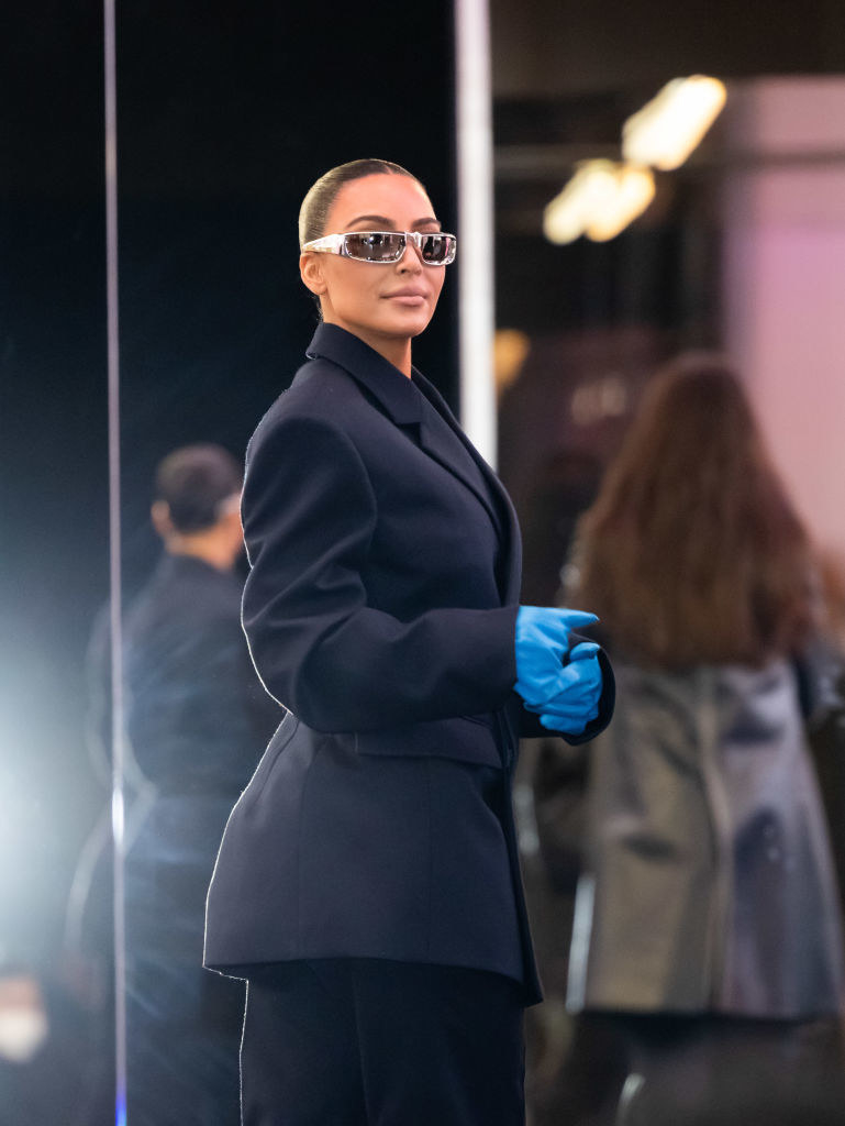 Kim wearing sunglasses and a short coat and gloves looks at the camera