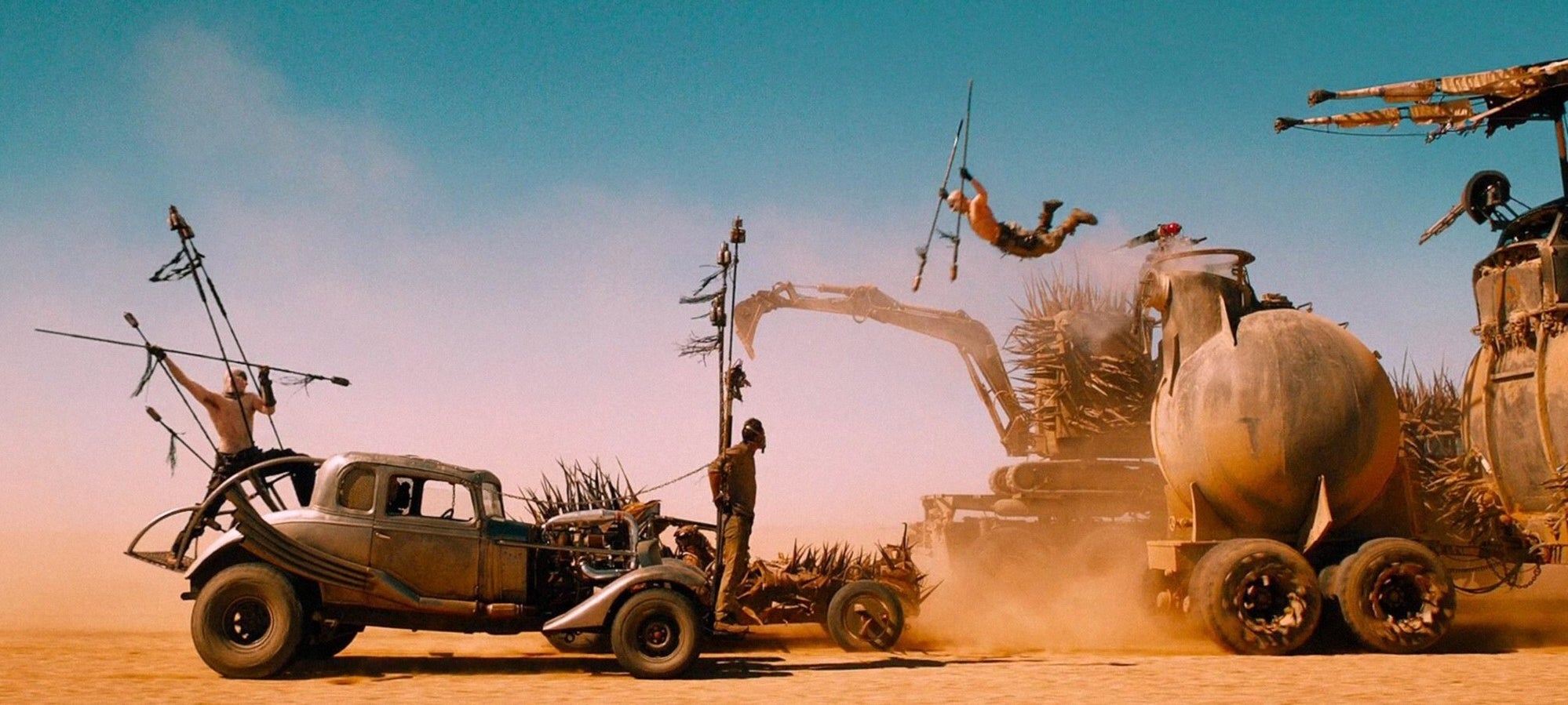 Heavily modified vehicles driving through a desert with people standing and jumping from them holding spears