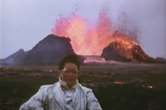Katia Kraft, who has short dark hair and thick rimmed glasses, standing with an erupting volcano behind her