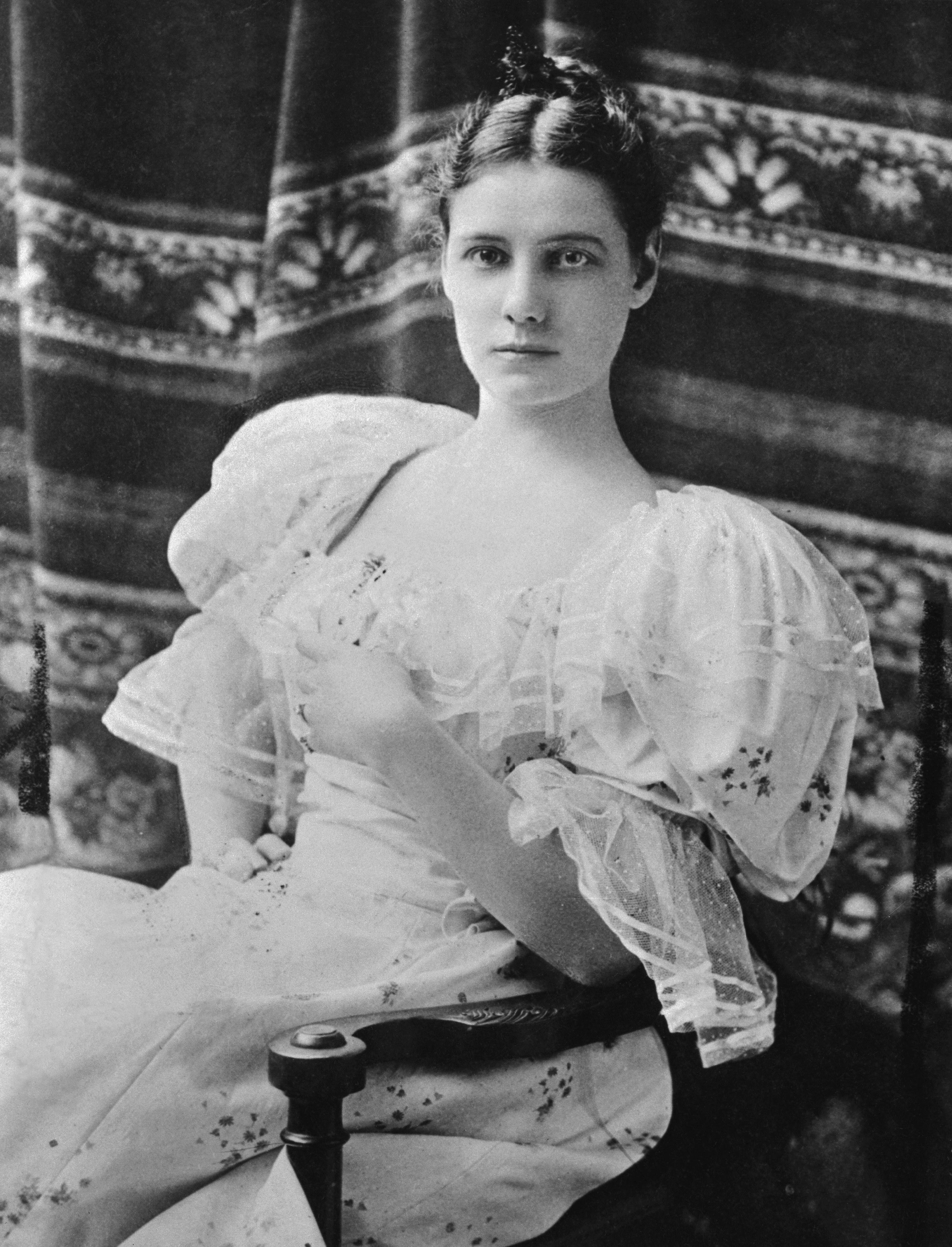 A formal portrait of Nellie Bly, with her hair tied back and wearing a ruffled dress as she sits on a chair and looks straight into the camera