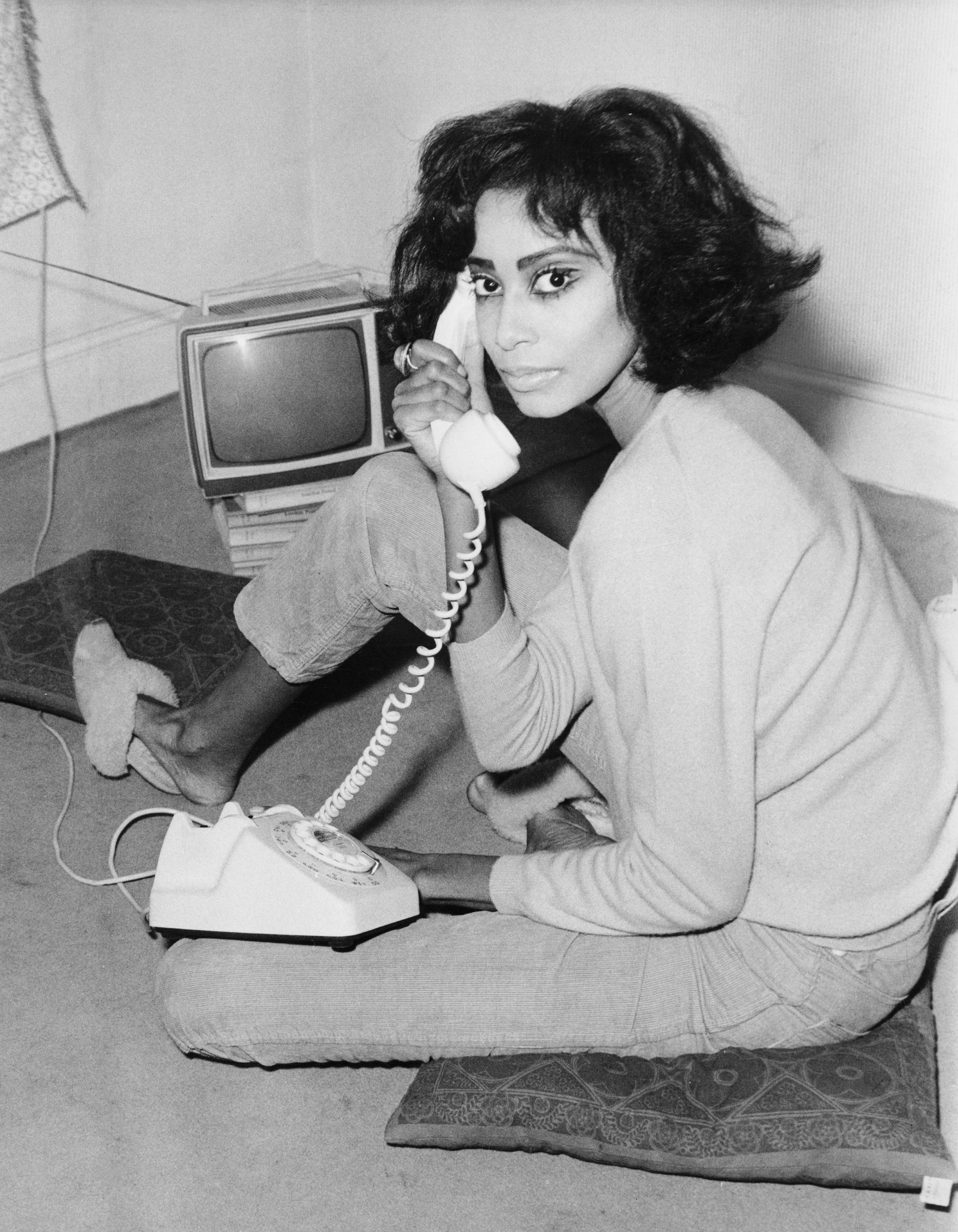Donyale Luna, wearing a long sleeved top and jeans, sitting on a cushion on the floor. She is holding a corded phone to her ear and looking into the camera with a TV in the background