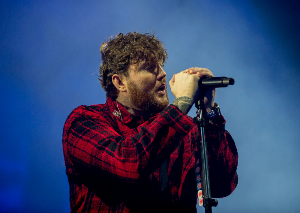 James Arthur in a red check shirt, holding a microphone in both hands while he sings