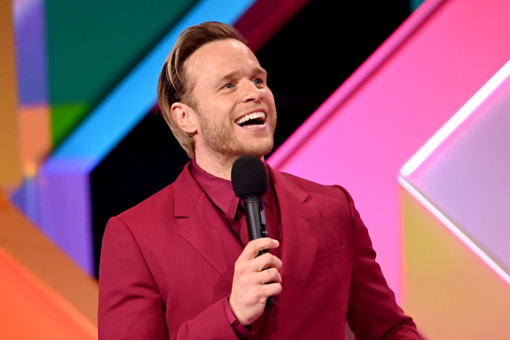 Olly Murs presenting at the Brit awards. He is wearing a red suit with a matching shirt and he&#x27;s holding a mic in his hand