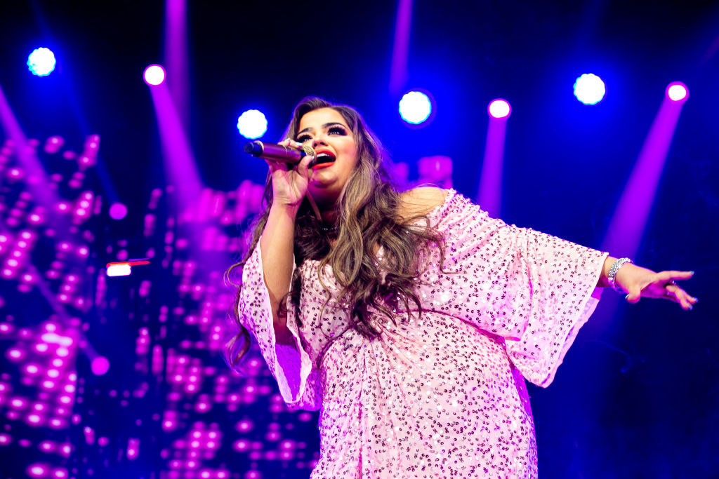 Scarlett Lee in a pink sparkly dress singing on stage. A mic is on her left hand while her right hand is out to the side