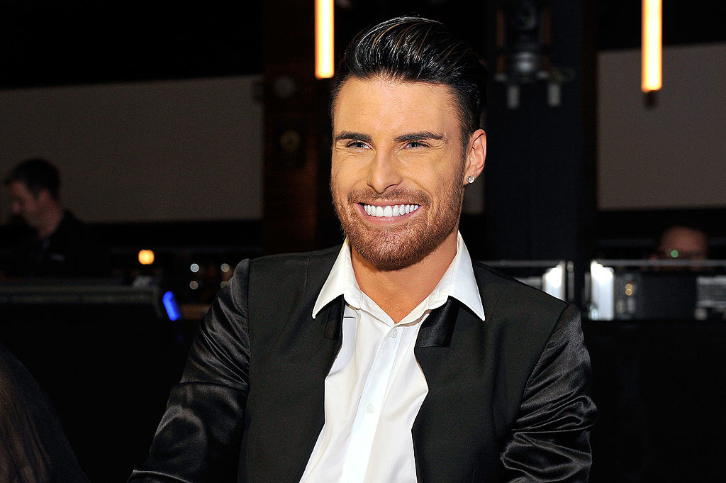 Rylan Clark in a black jacket and white shirt, sat down smiling