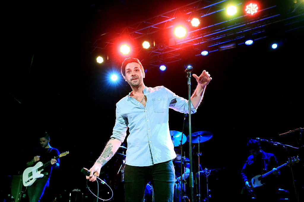 Ben Haenow on stage, standing in front of a band, in black jeans and a shirt. He has a microphone in one hand while holding the stand in the other