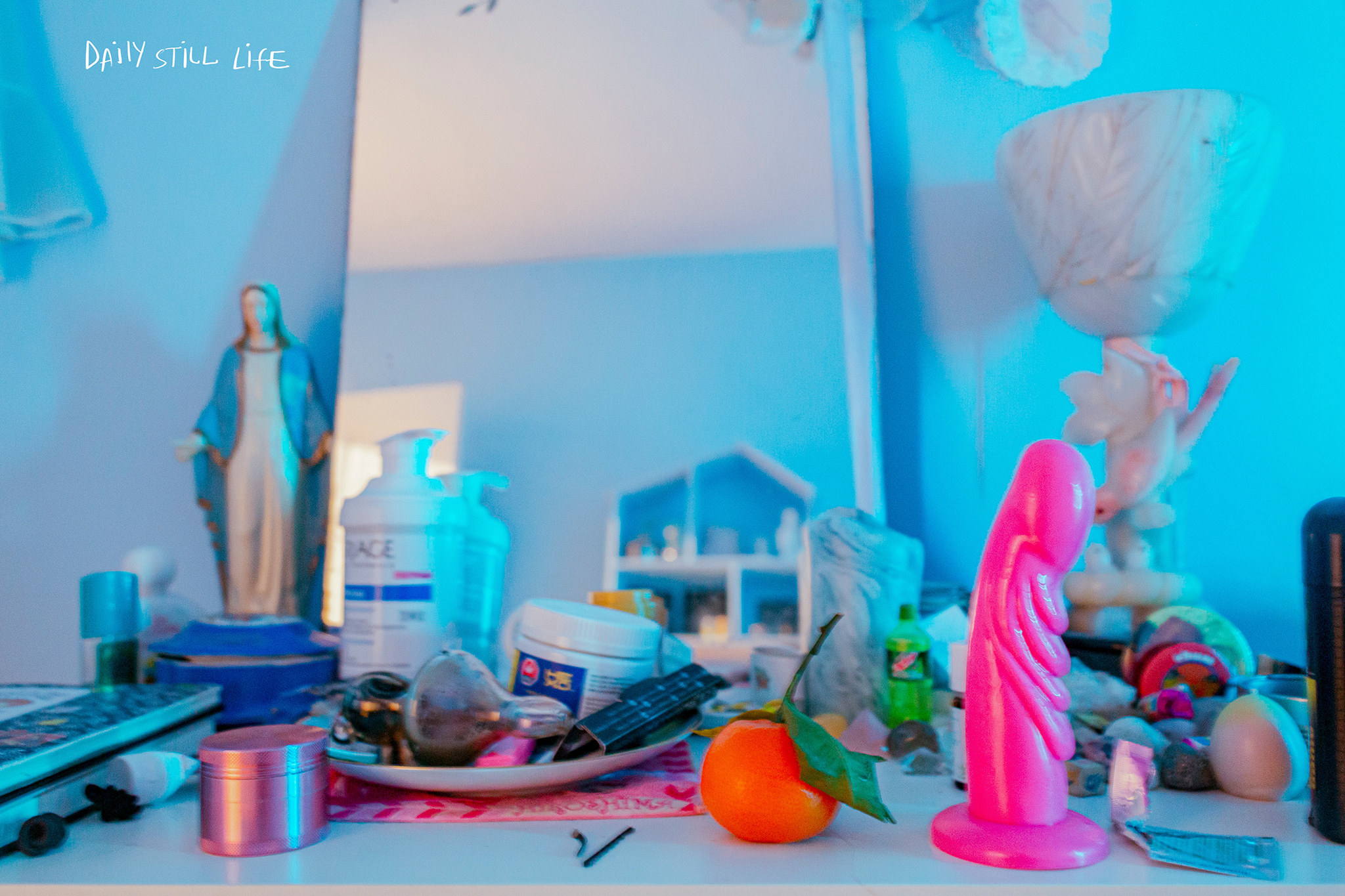 A dresser top with a pink dildo, a statue of mary, an orange, and other paraphenalia. 