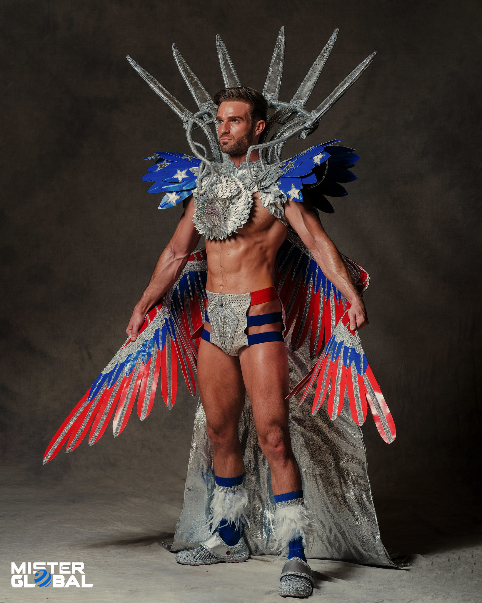 A bare-chested man wears an outfit with the colors of the US flag, including a winged shoulder piece with a wide, Statue of Liberty crown and three-strap loincloth