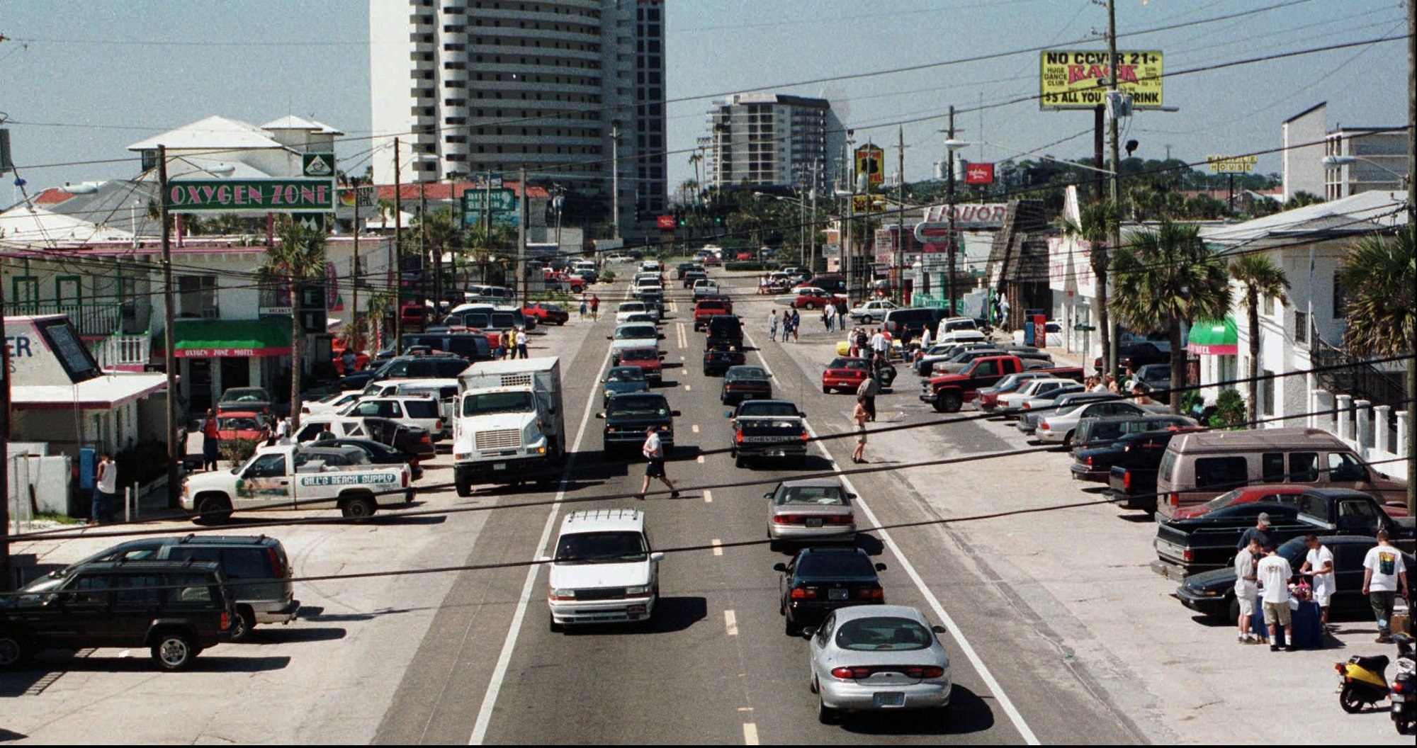 A road with cars and people walking that is a main drag with buildings around it 