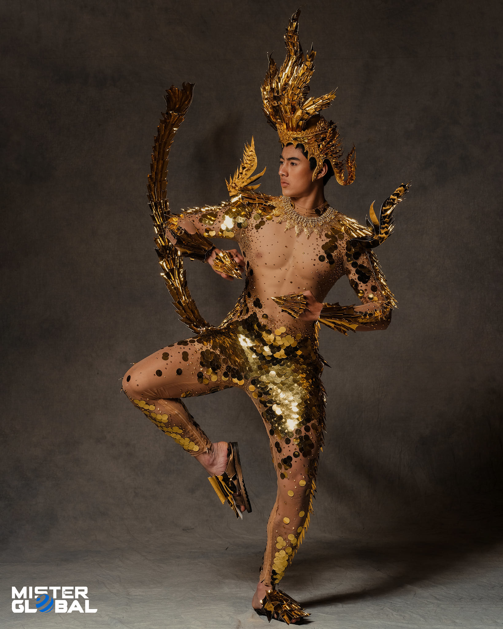 Man balanced on one leg and wearing an ornate headdress and tight, bare-chested bodysuit with metallic-looking sequins