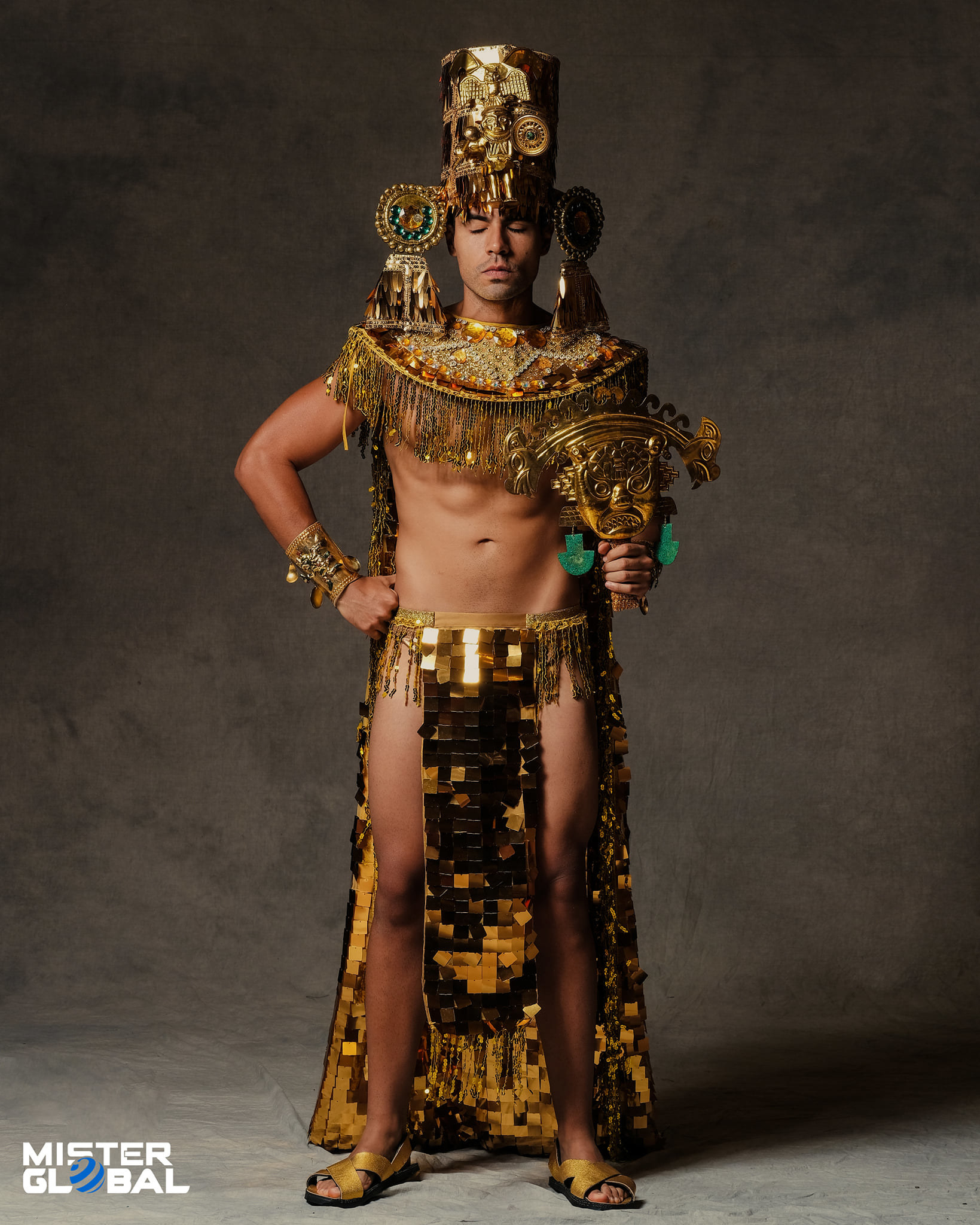 Man with eyes closed wearing ornate gold headdress, chest-baring gold outfit, and sandals