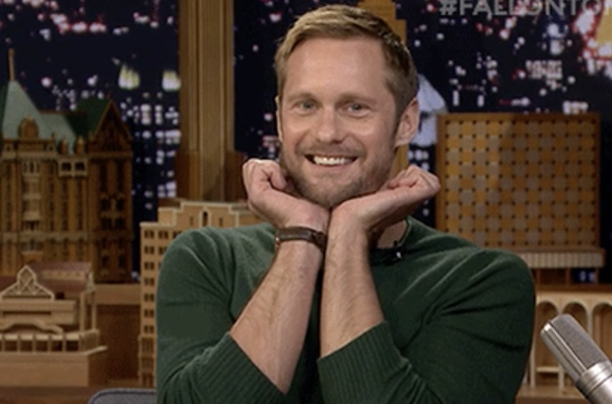 alexander skarsgard with his hands under his chin playfully smiling
